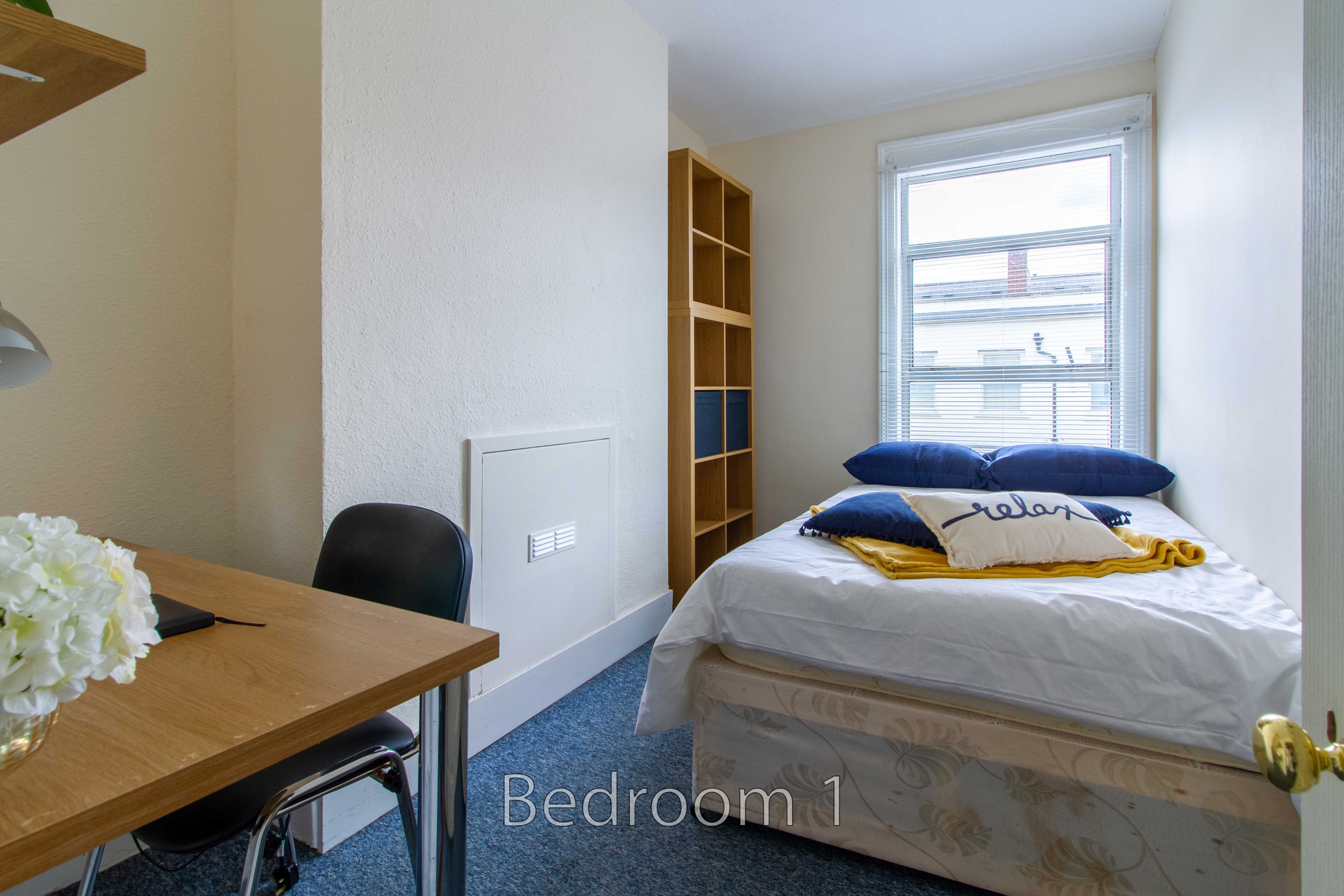 A furnished bedroom with a bed, desk, shelving unit, window and blue carpet.