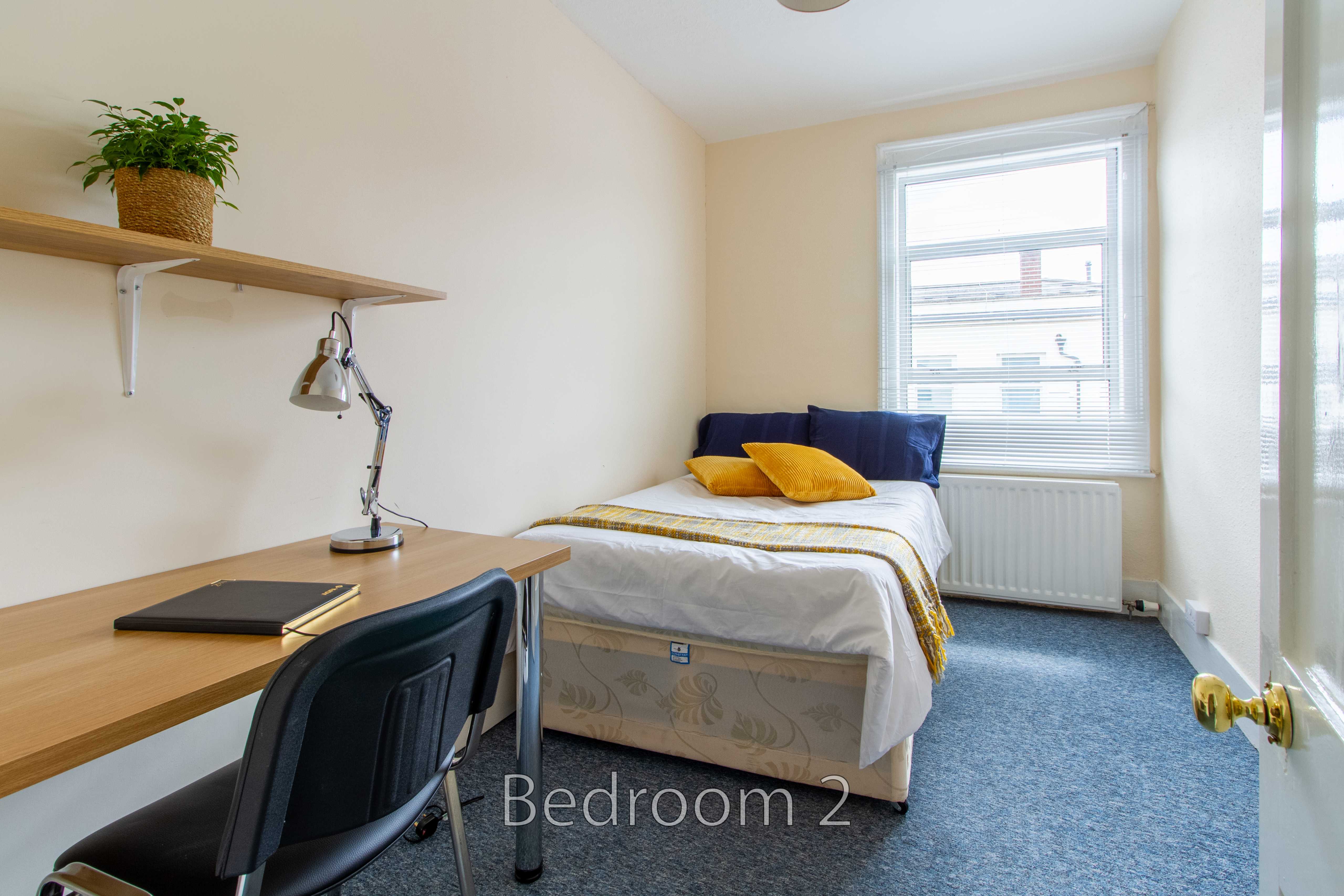 A small bedroom with a single bed, desk, chair, wall shelf, window and radiator.