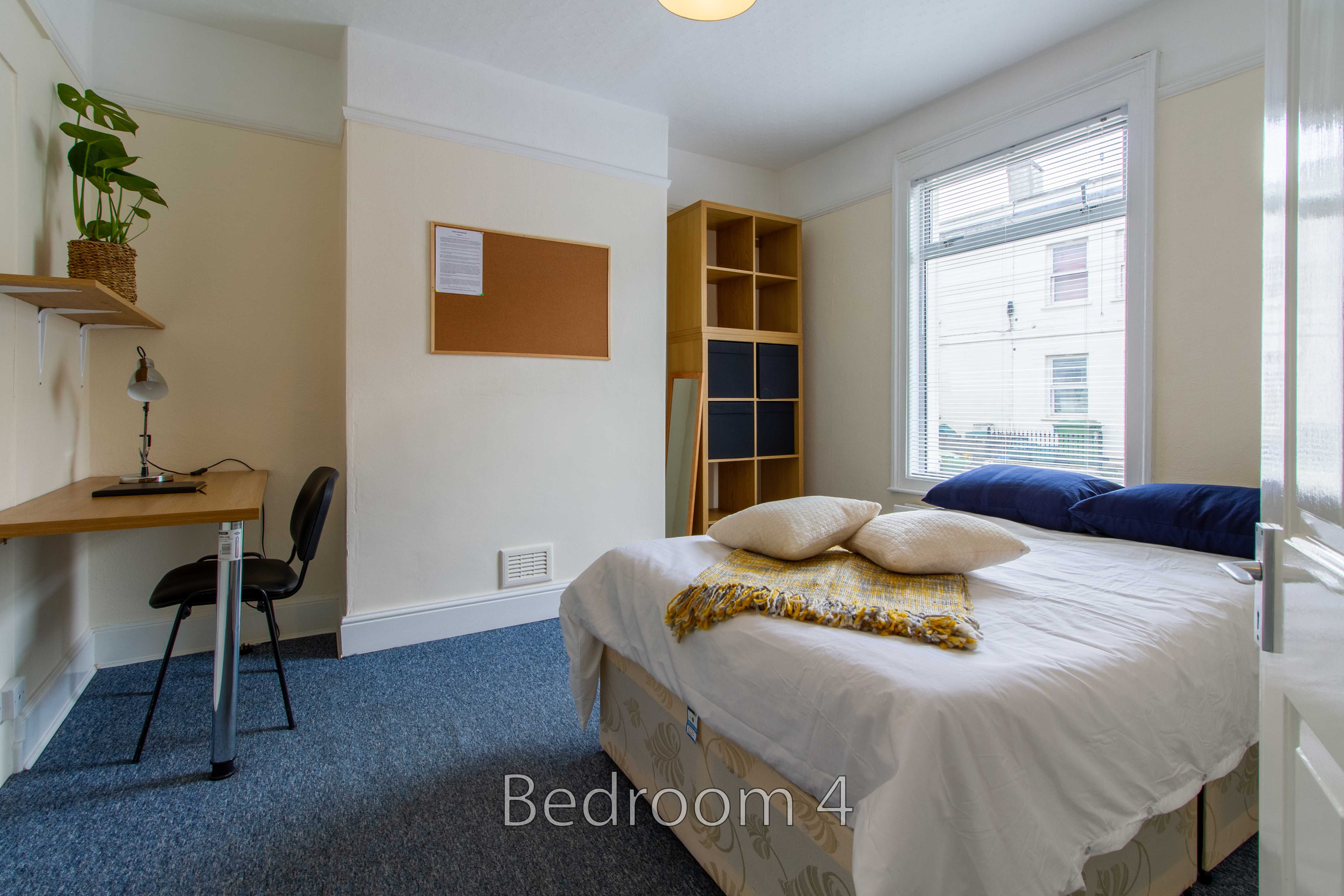A furnished bedroom with a bed, desk, shelving, window, and blue carpet.