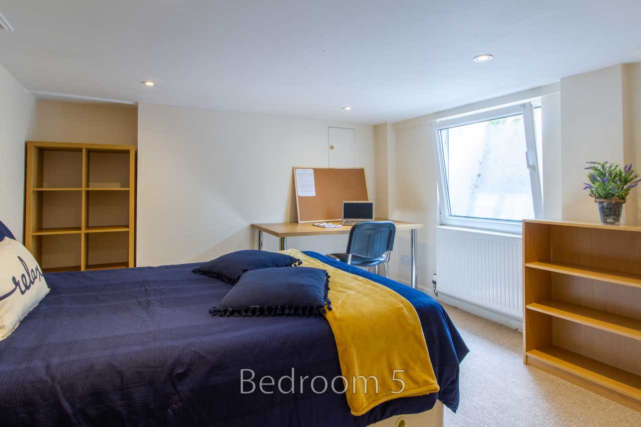A furnished bedroom with a double bed, desk, chair, shelving and a window.
