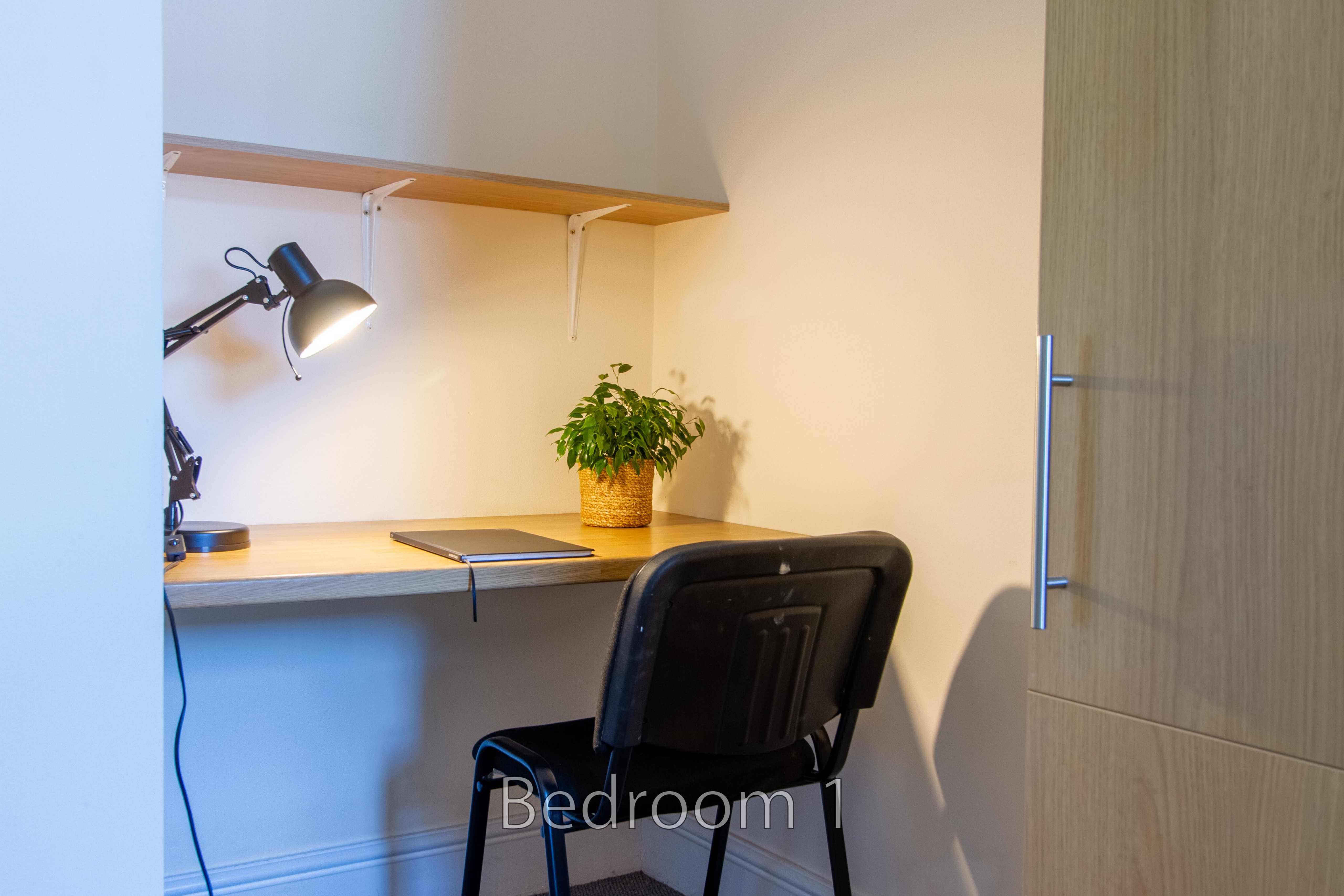 A compact bedroom workspace with a desk, chair, lamp, shelves and a potted plant.