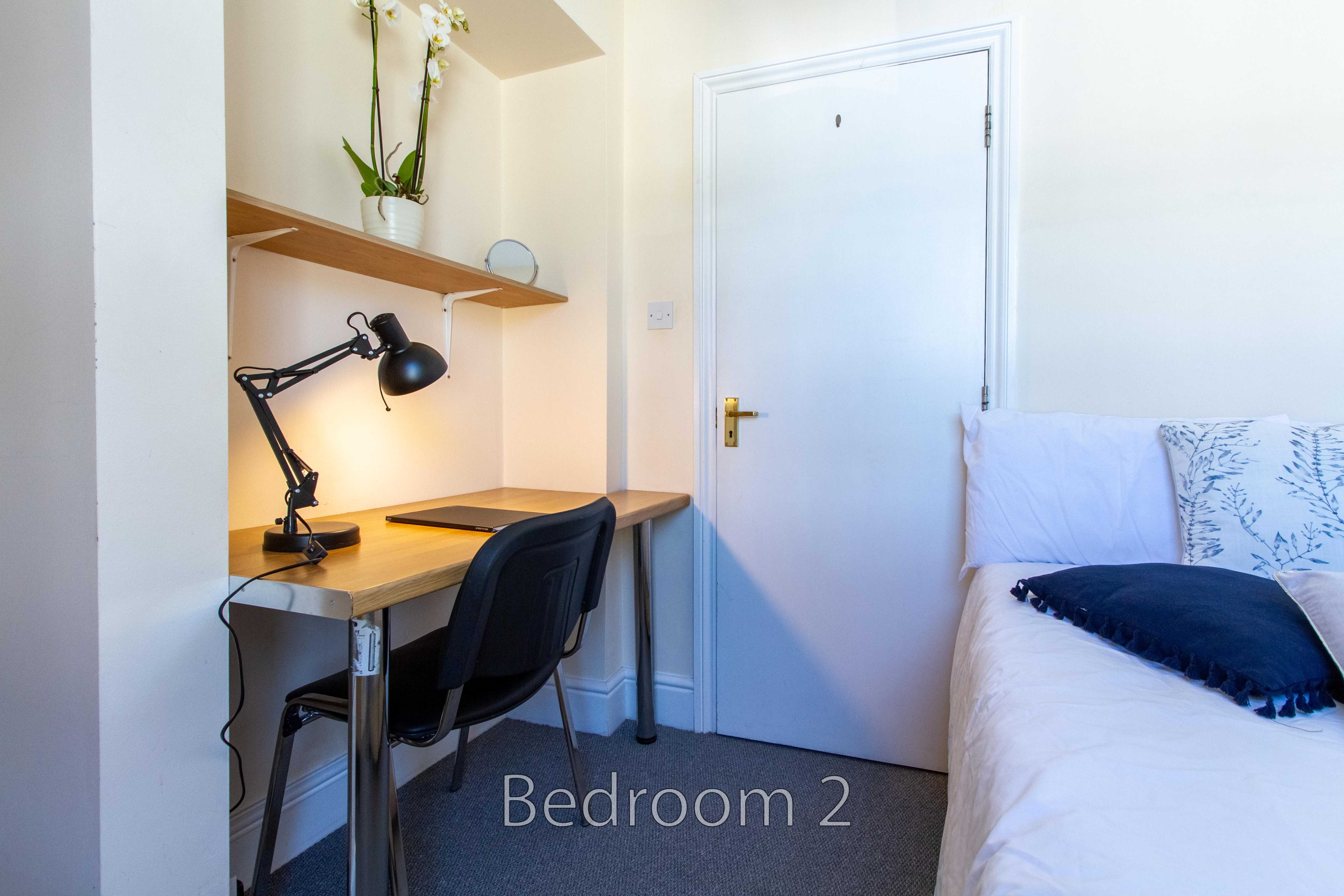 Bedroom with a single bed, desk, chair, lamp and wall shelves.