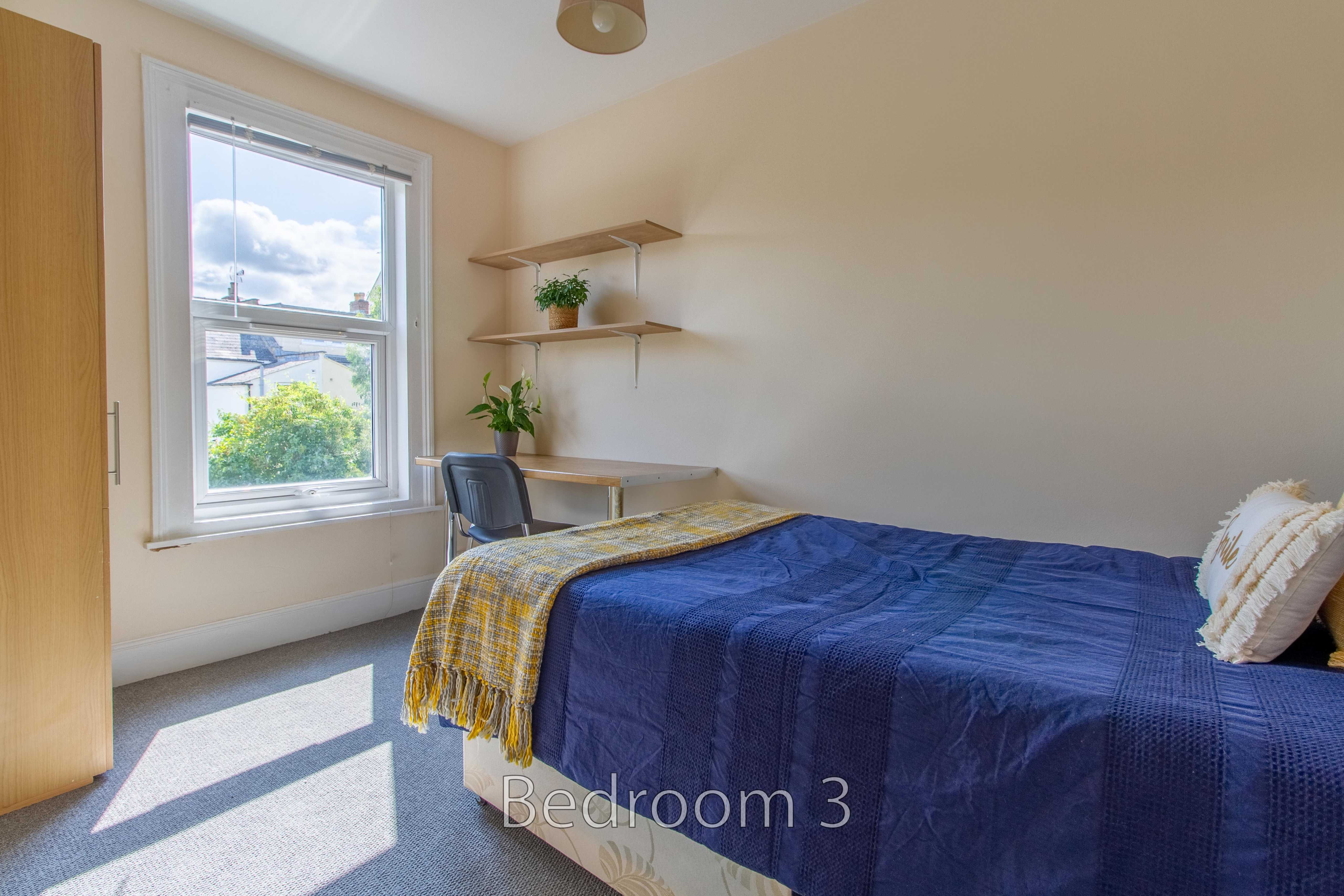 A bright bedroom with a bed, desk, shelving, wardrobe, window and blue bedspread.