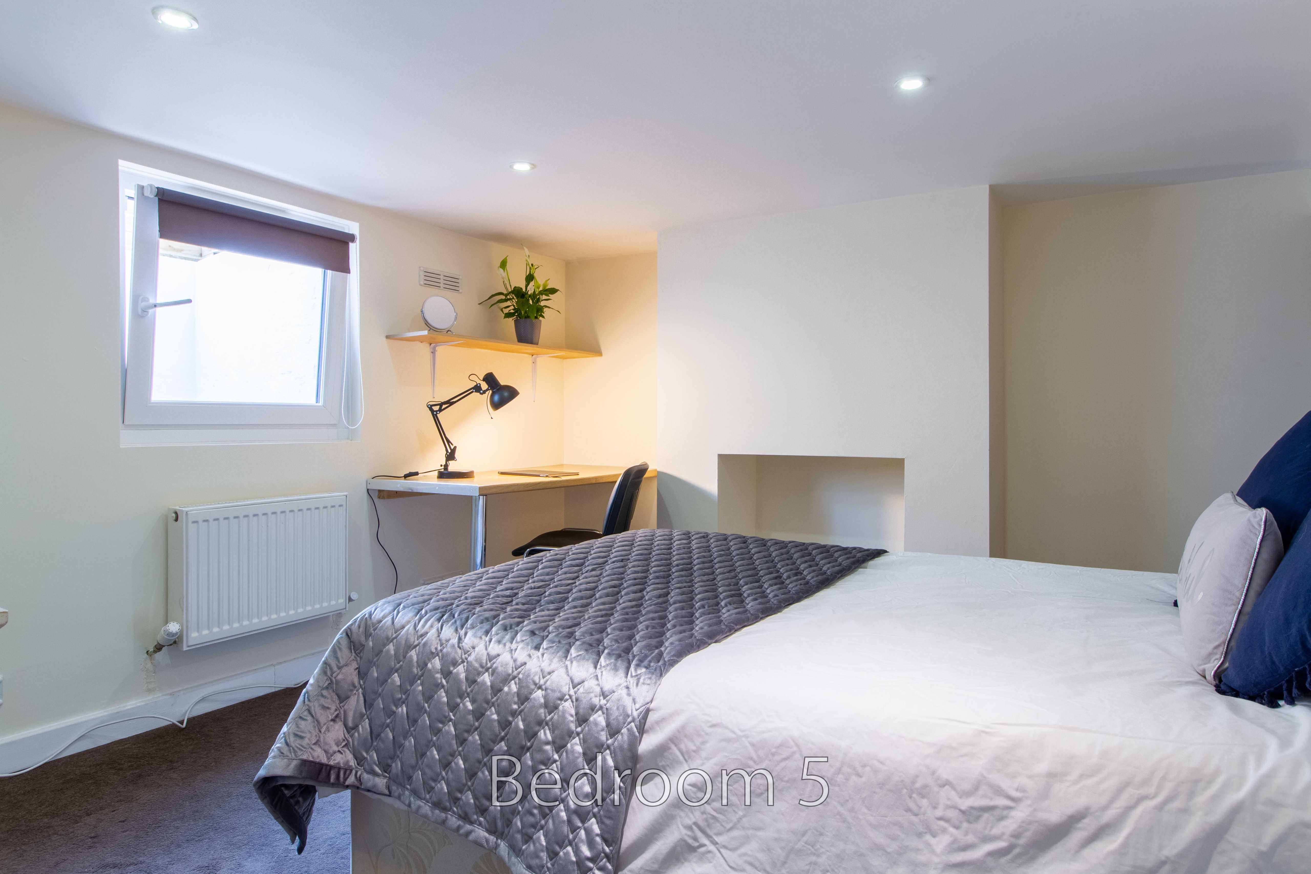 A bedroom with a bed, desk, chair, window, radiator and wall shelf.