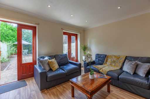 A furnished living room with sofas, a wooden coffee table, recessed ceiling lights, and glazed doors opening to a garden.