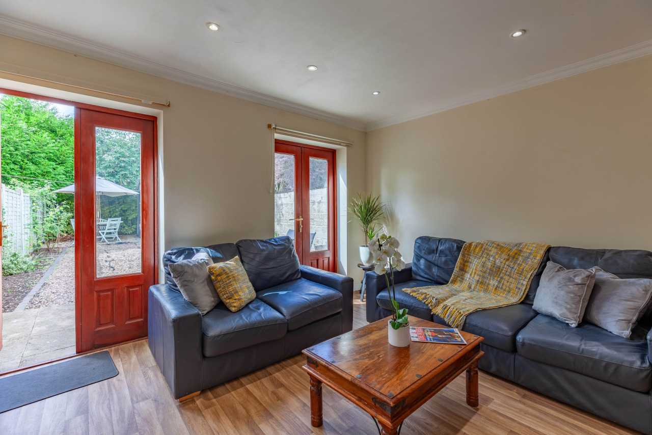 A furnished living room with sofas, a wooden coffee table, recessed ceiling lights, and glazed doors opening to a garden.