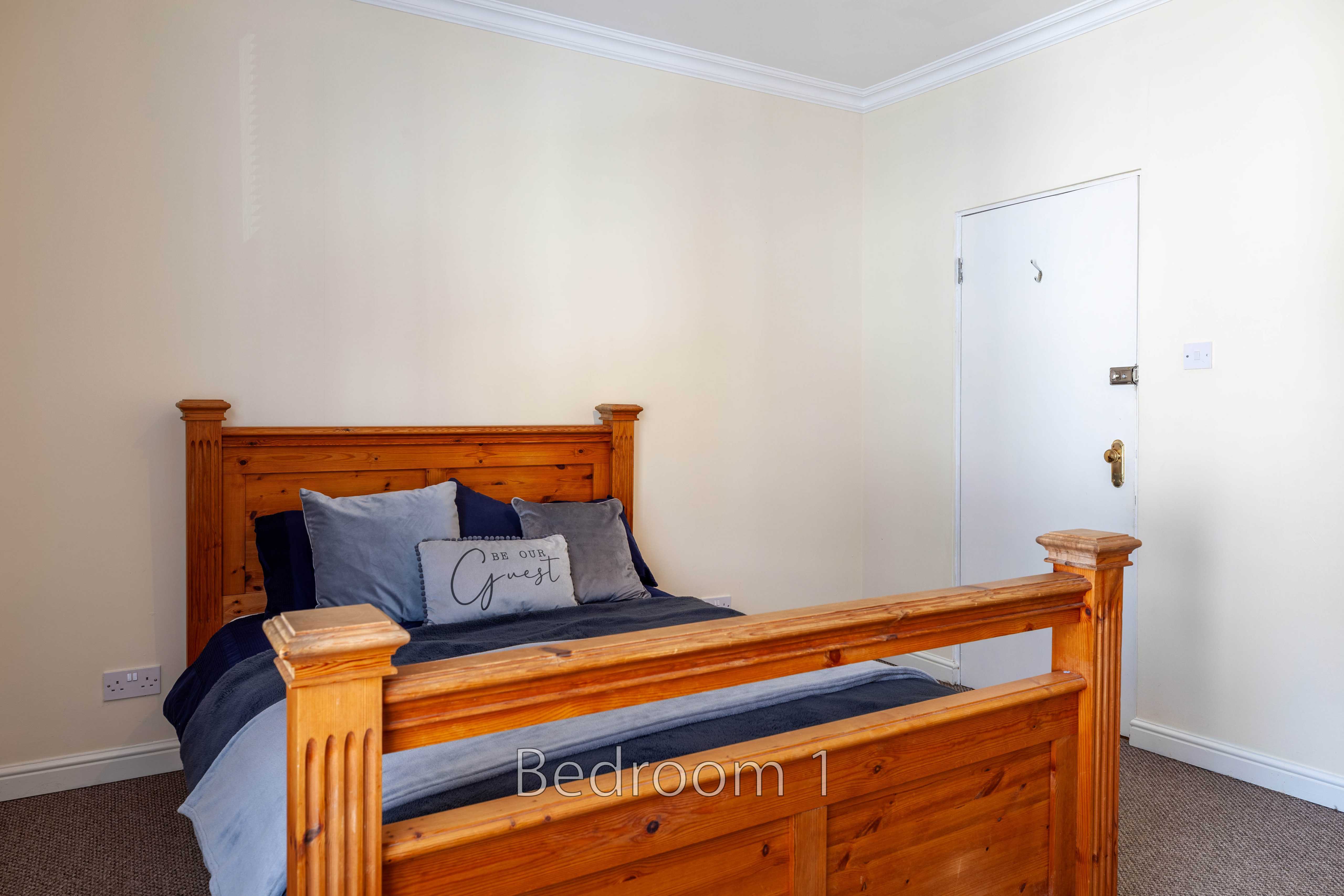 A furnished bedroom with a wooden bed, blue bedding, cream walls and a white door.