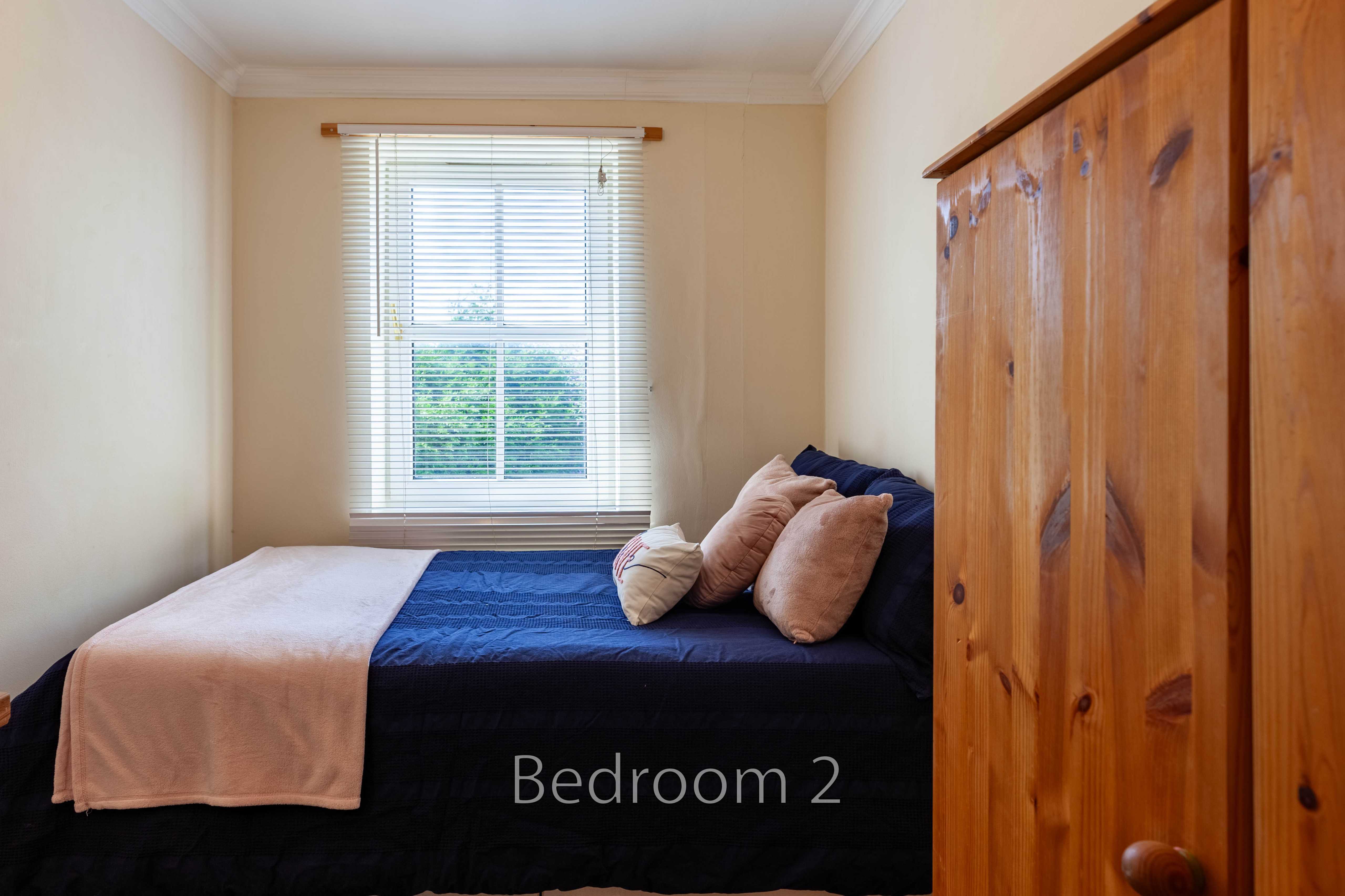 A bedroom with a double bed, pillows, a window with blinds, and a large wooden wardrobe.