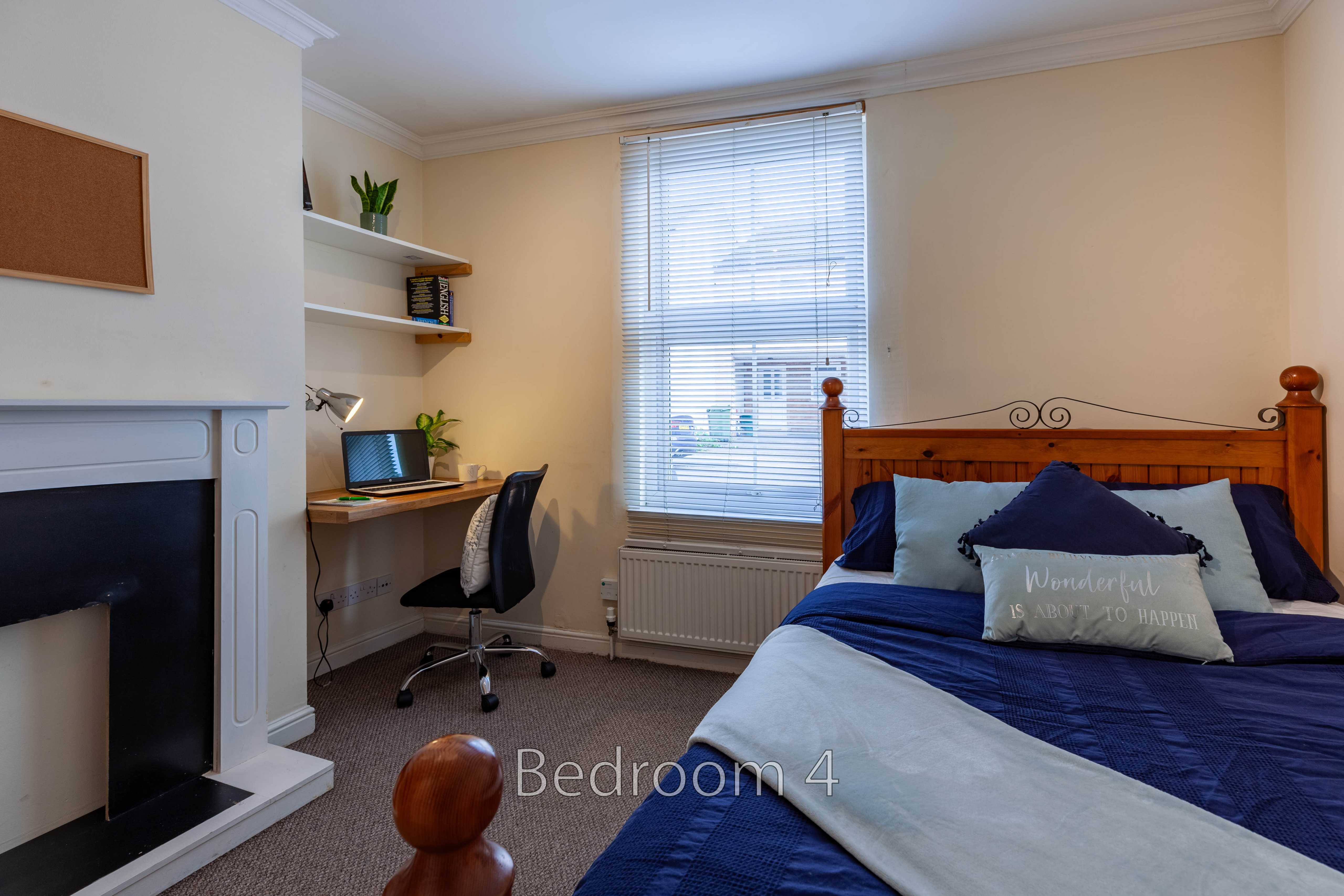 A bedroom with a wooden bed, blue bedding, a desk and chair, shelving, a window, and a fireplace.