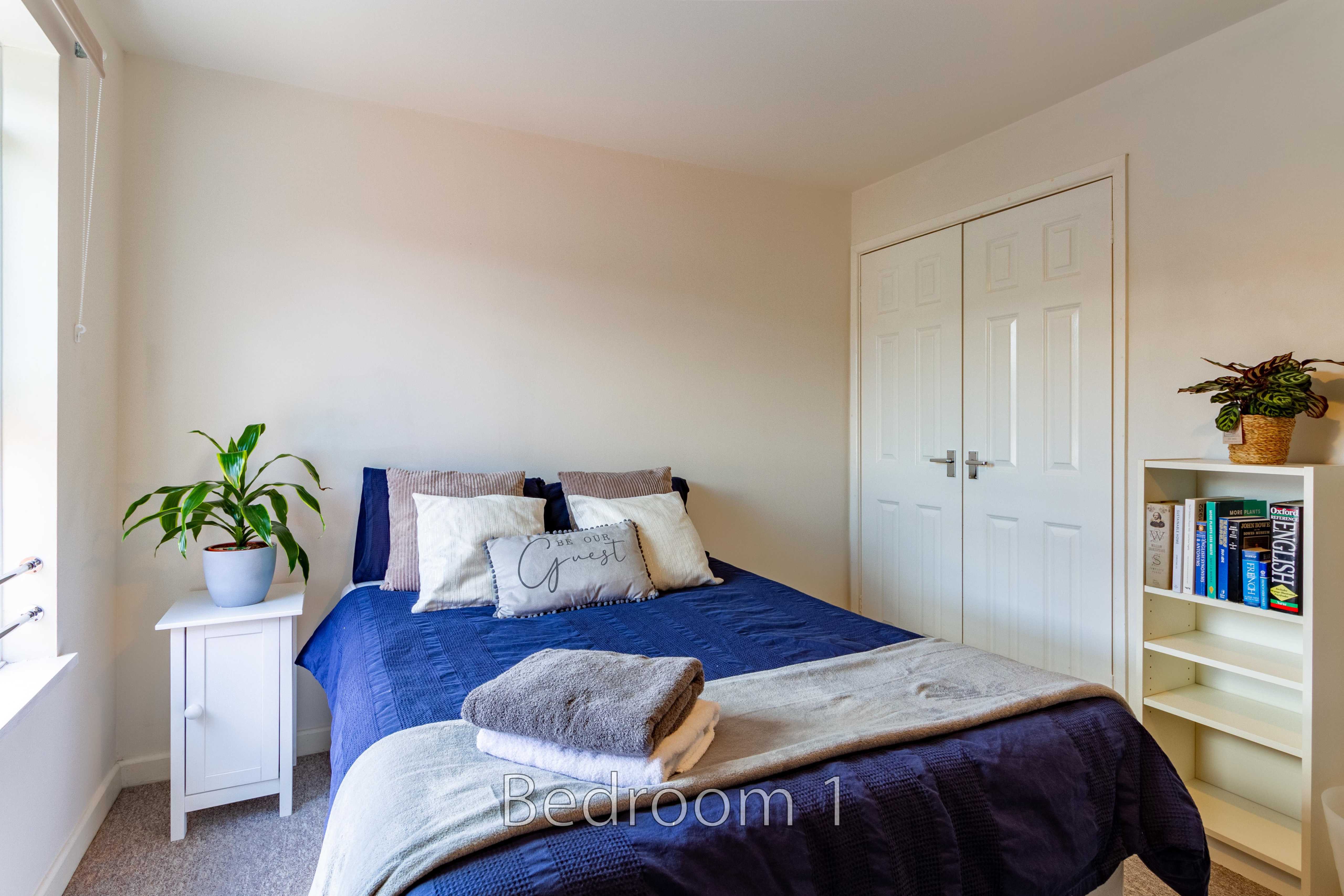 A furnished bedroom with a double bed, white bedside table, plants, bookshelf and fitted wardrobe.