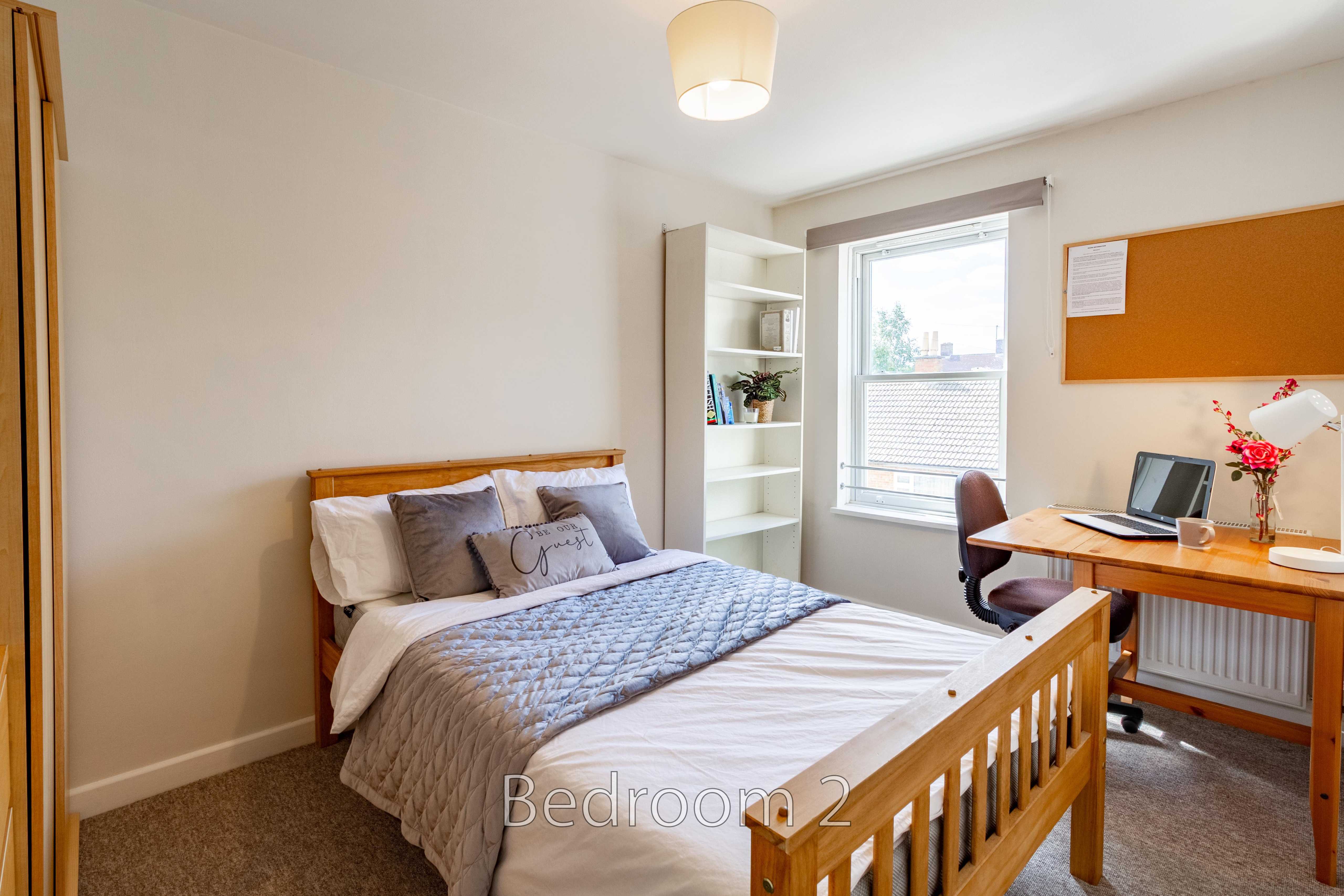 A furnished bedroom with a wooden bed, desk, chair, shelving, window and noticeboard.