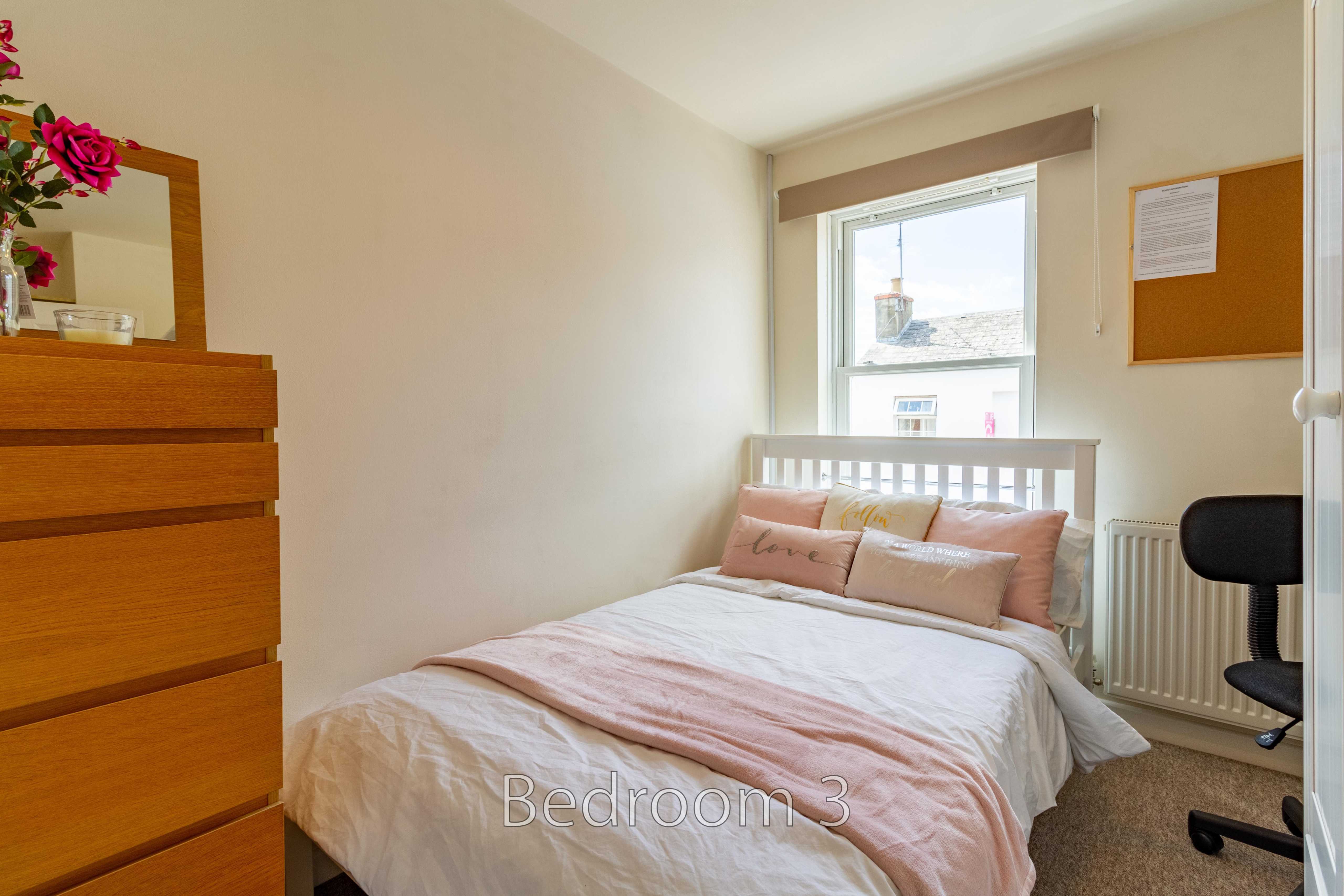 A small bedroom with a double bed, dresser, window, radiator and desk chair.
