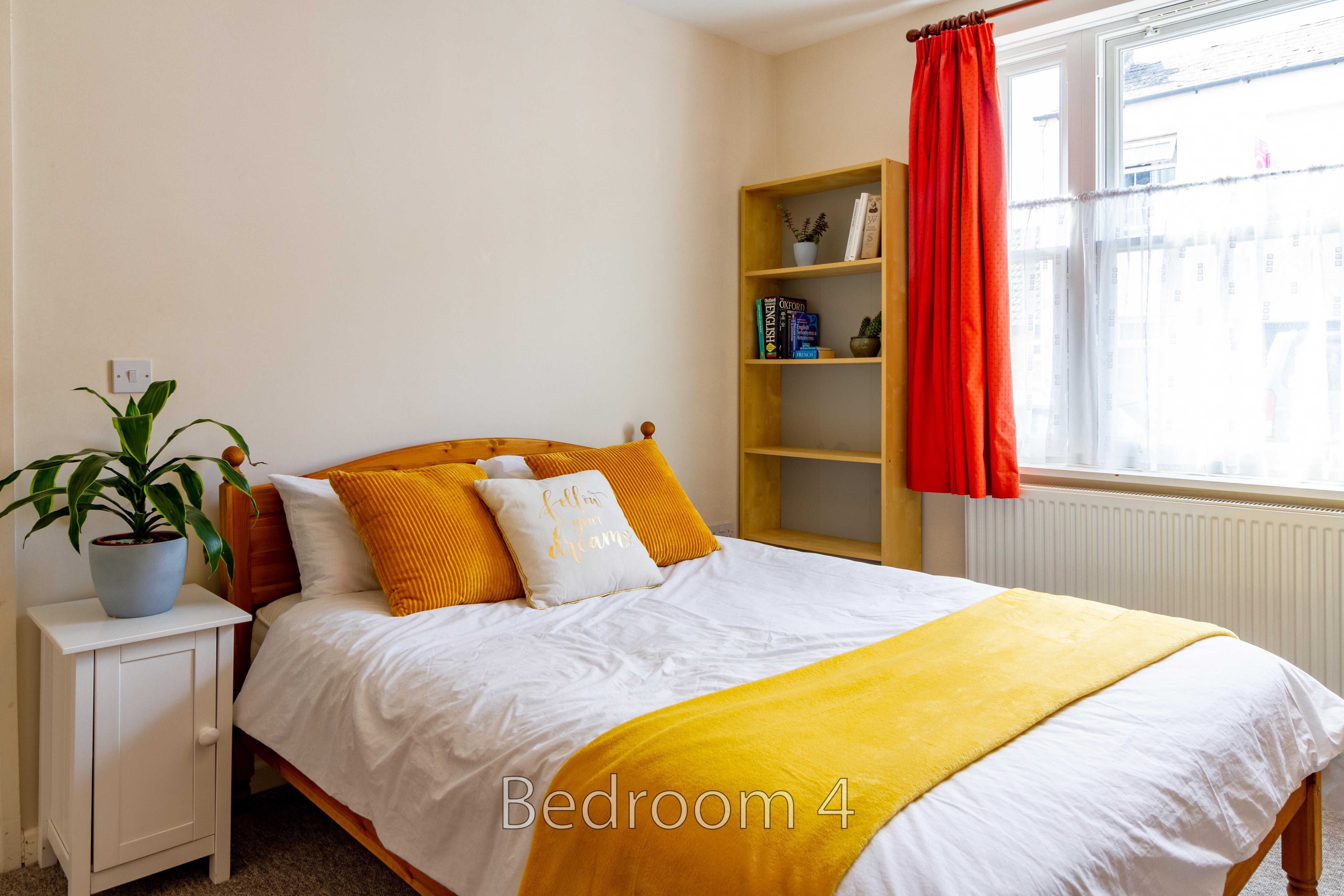 A bedroom with a double bed, yellow and white bedding, a bedside table, shelving, a plant and a window with red curtains.