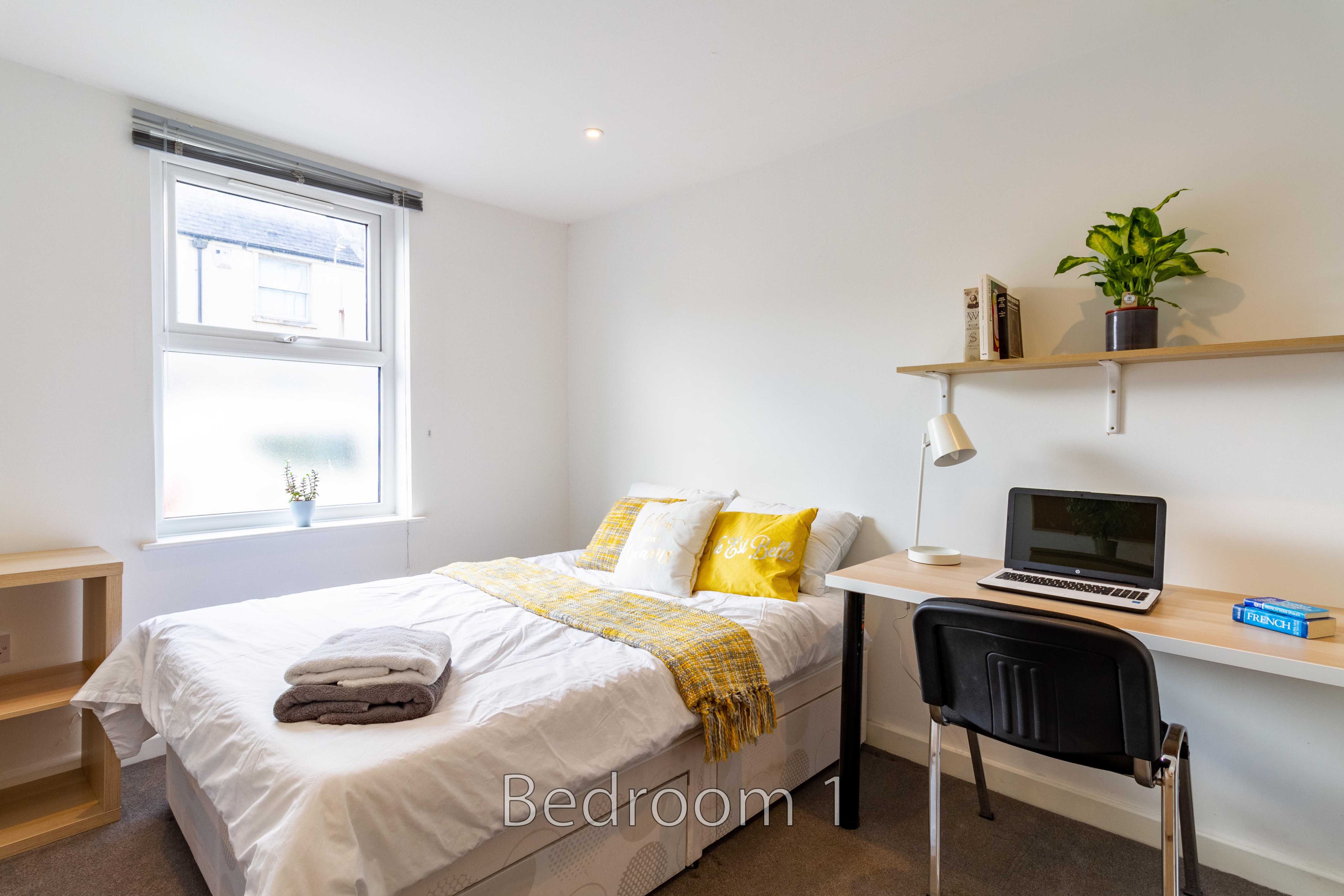 A bright bedroom with a double bed, desk, chair, window and wall shelf.