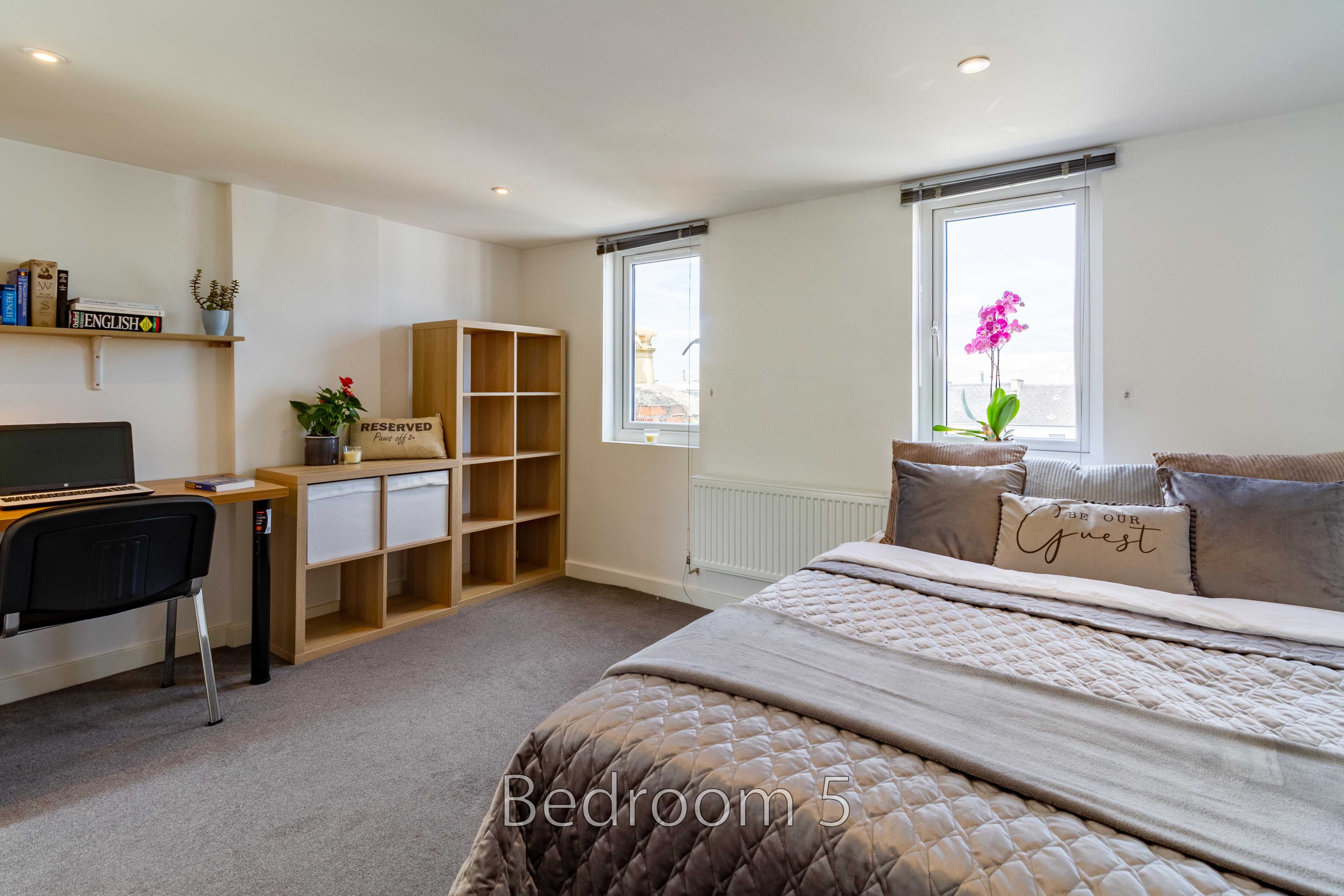 A furnished bedroom with a bed, desk, shelving, two windows and carpeted flooring.