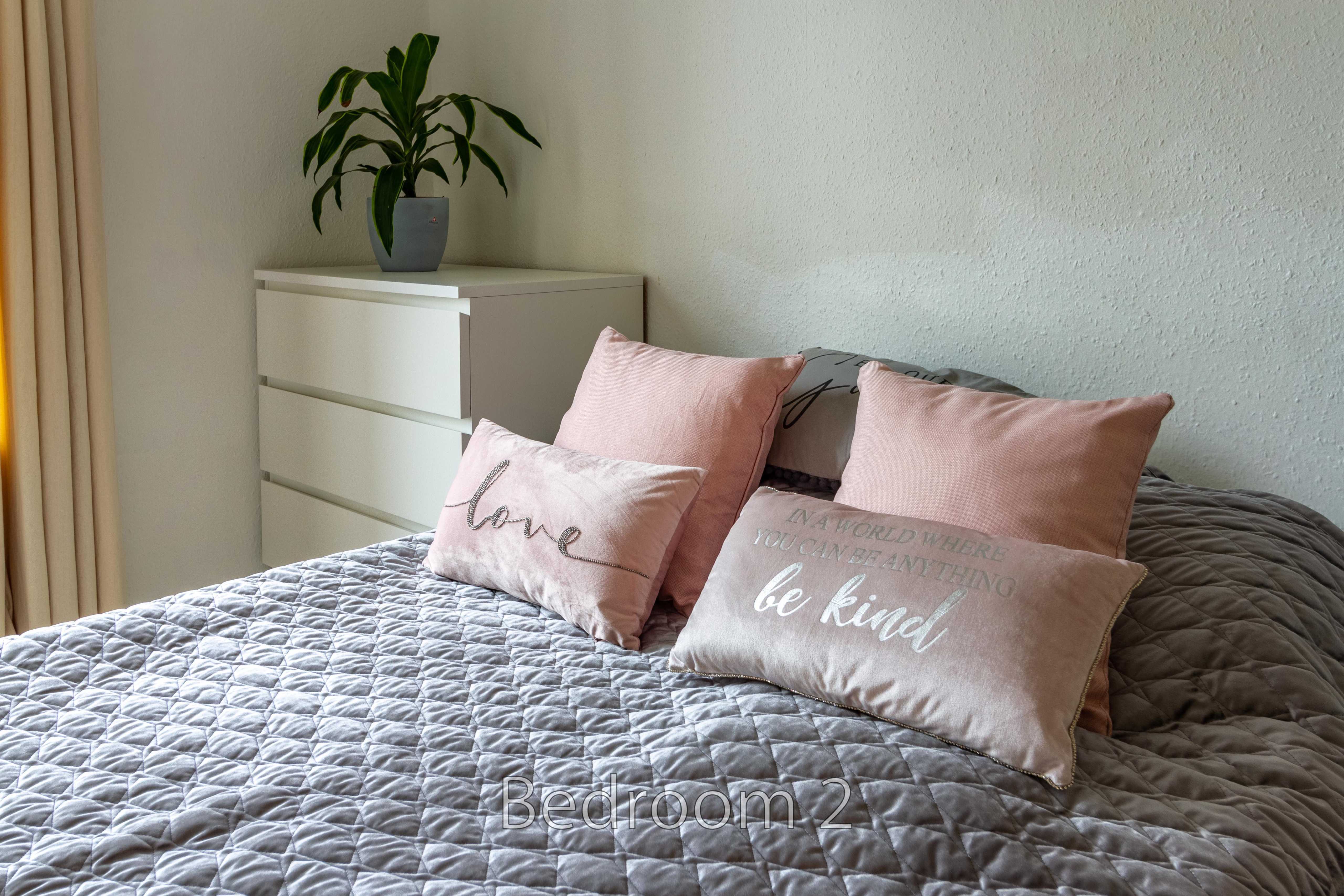 A bedroom with a neatly made bed, pink cushions, a white chest of drawers and a potted plant.
