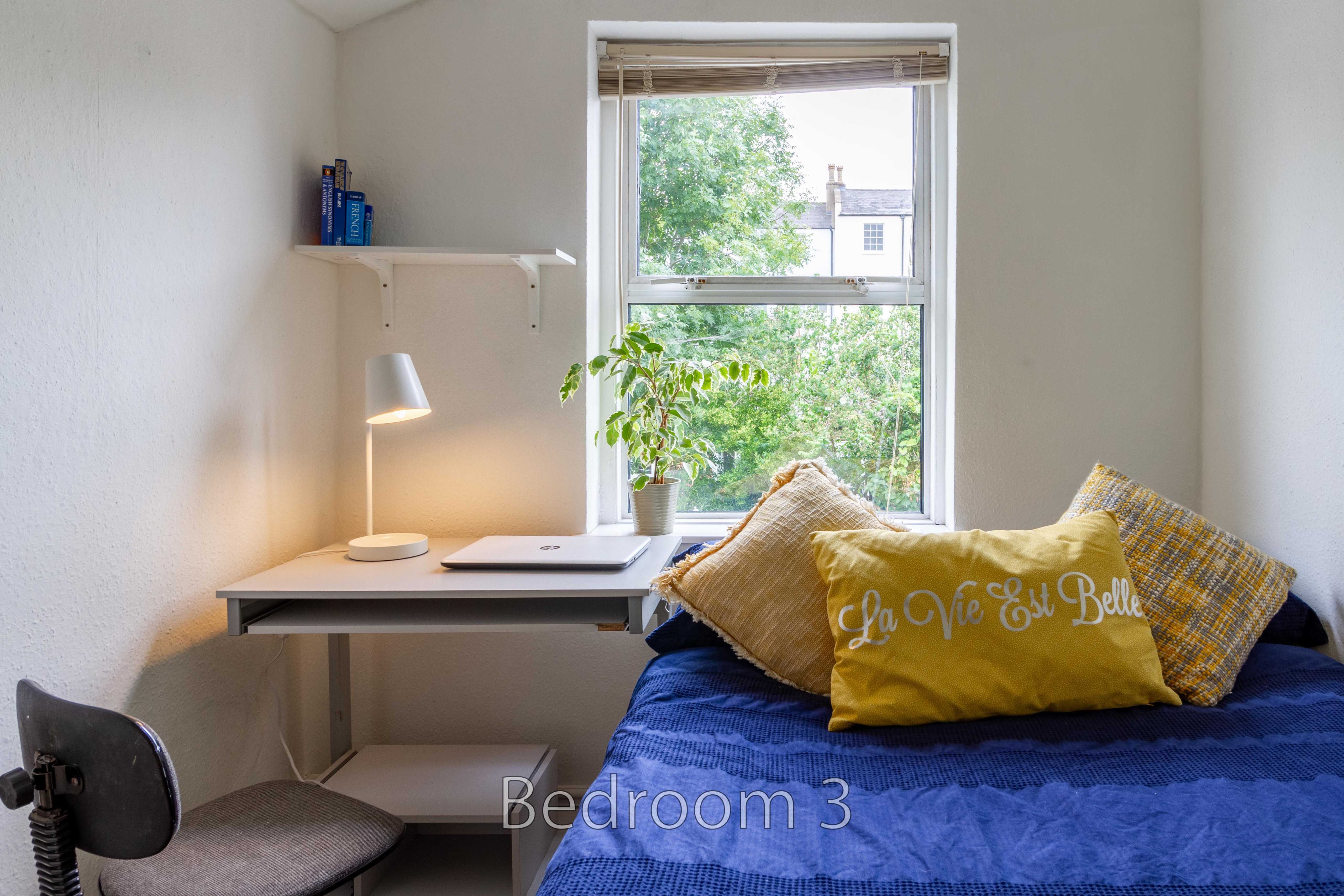 A small bedroom with a single bed, desk, chair, lamp and window.