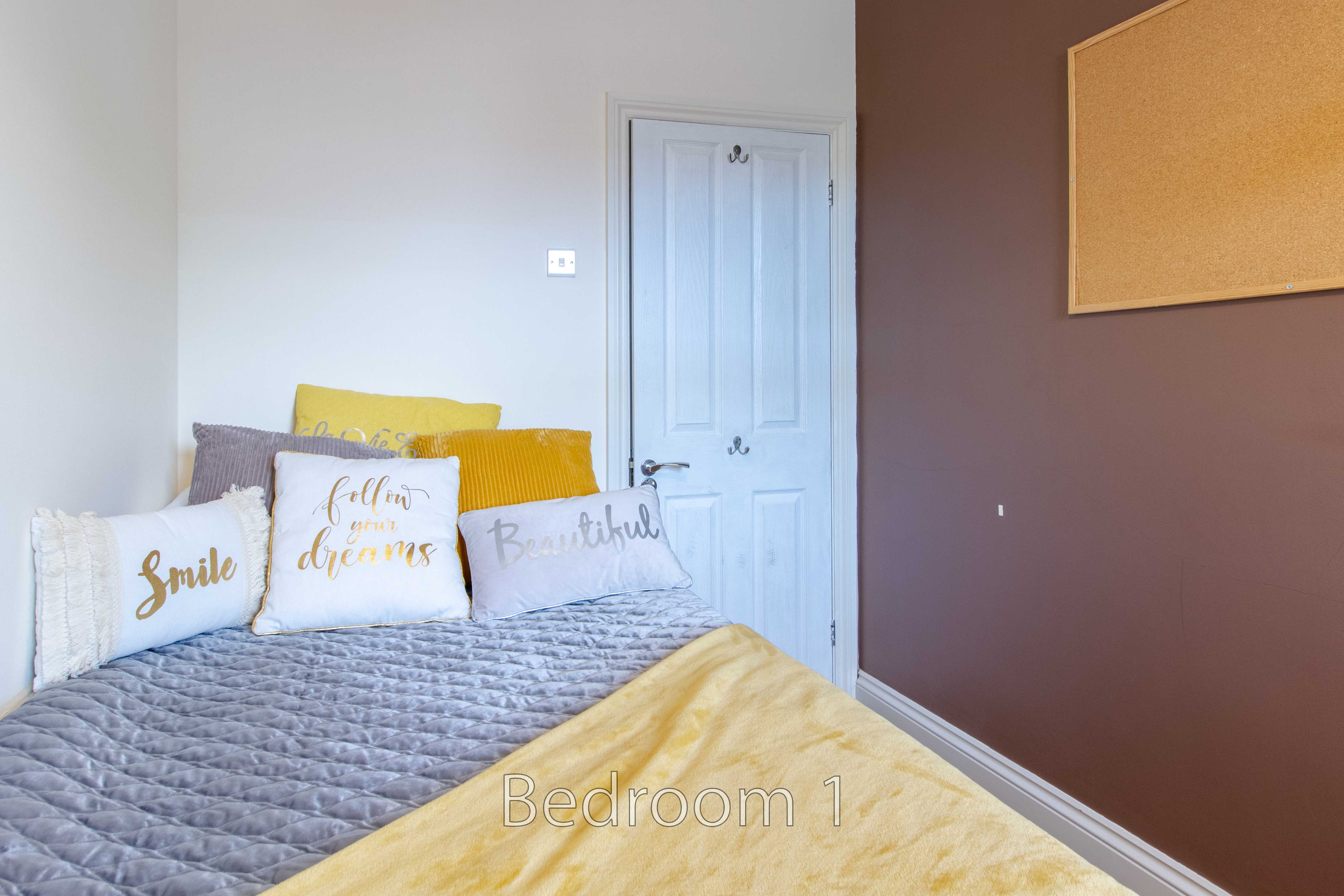 A bedroom with a neatly made bed, decorative pillows, a white door and a cork noticeboard on a brown wall.