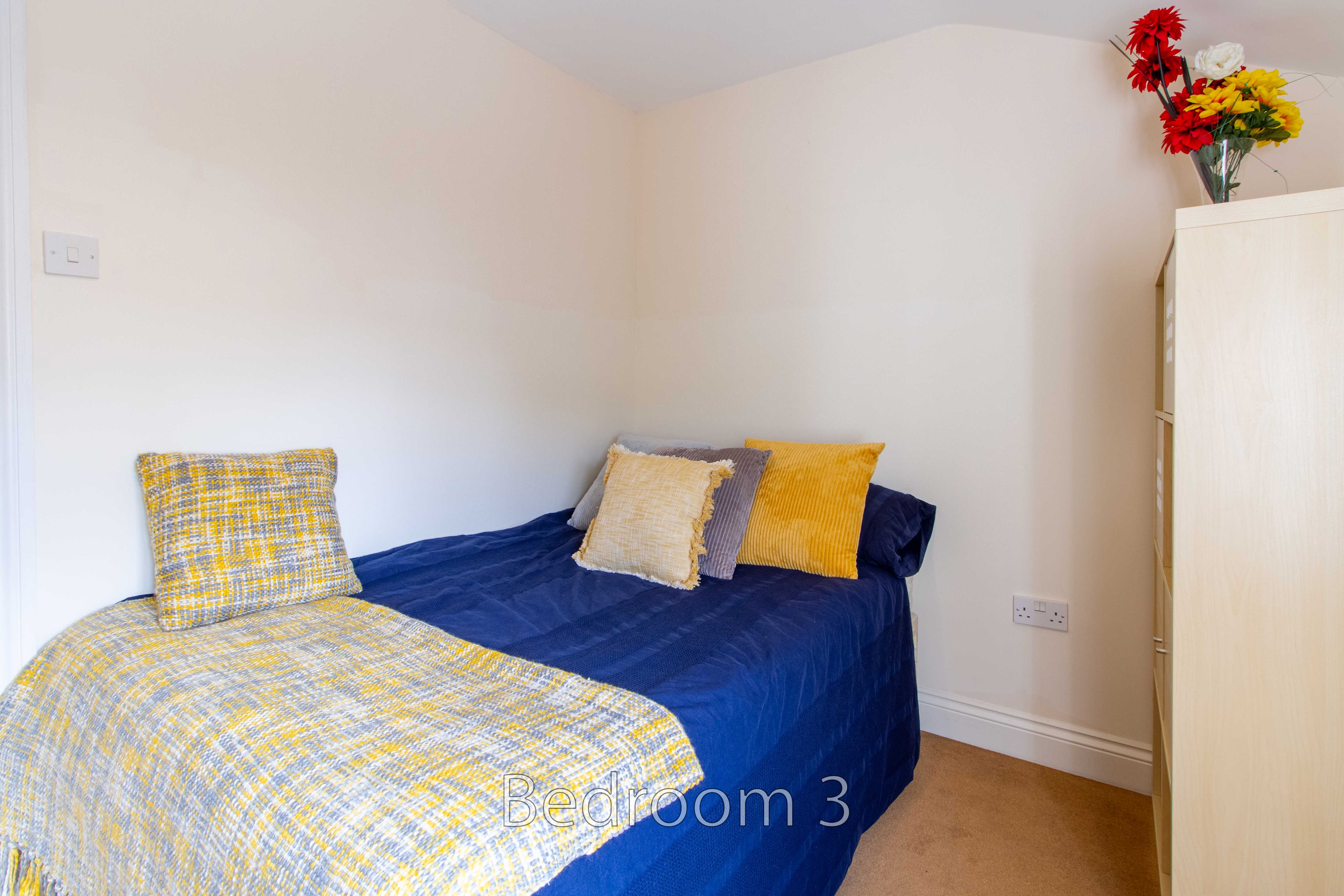 A furnished bedroom with a single bed, patterned blue and yellow bedding, a wardrobe, and a vase of flowers.
