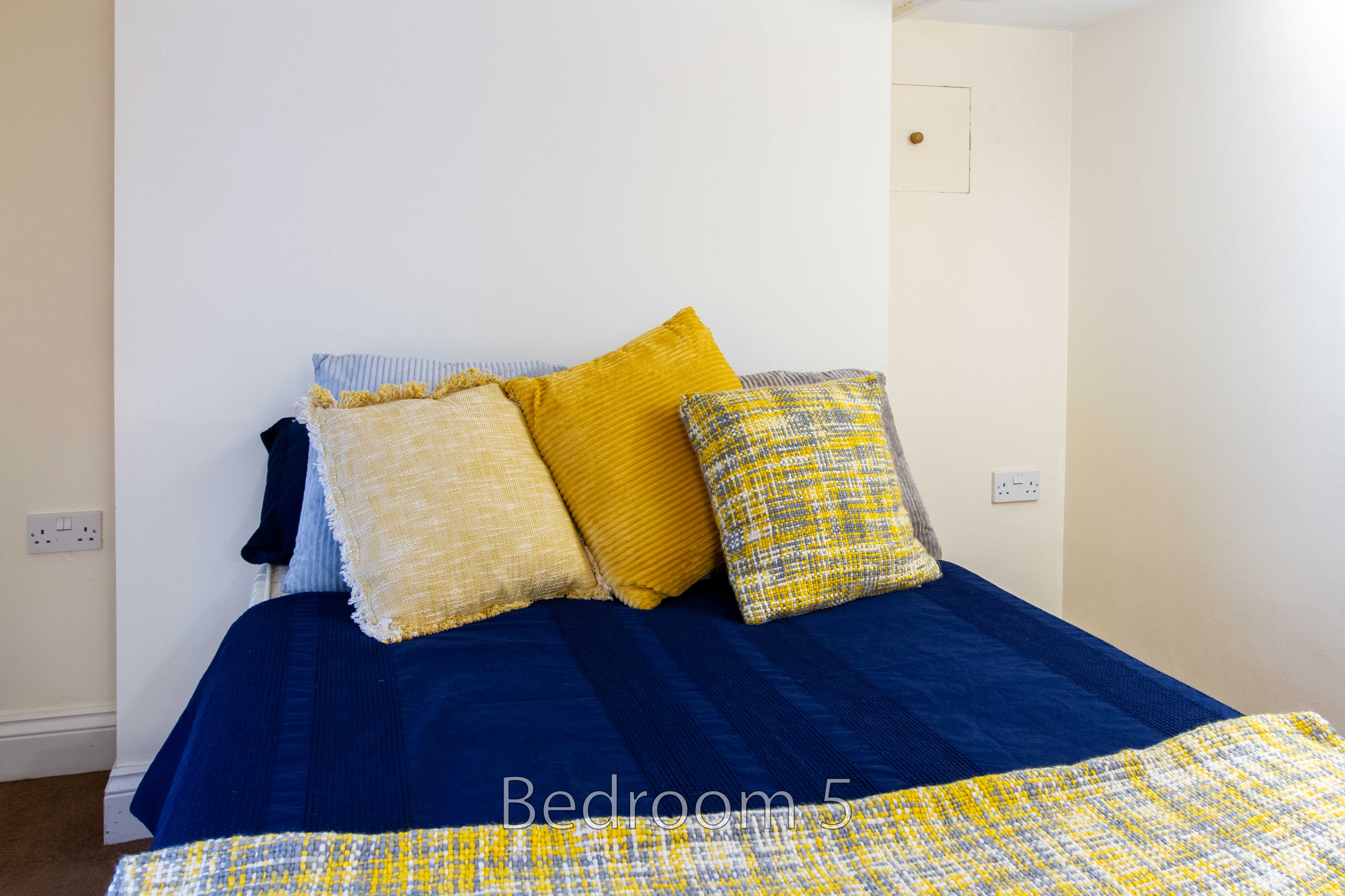 A bed with dark blue bedding and several yellow and patterned pillows in a light-colored room.