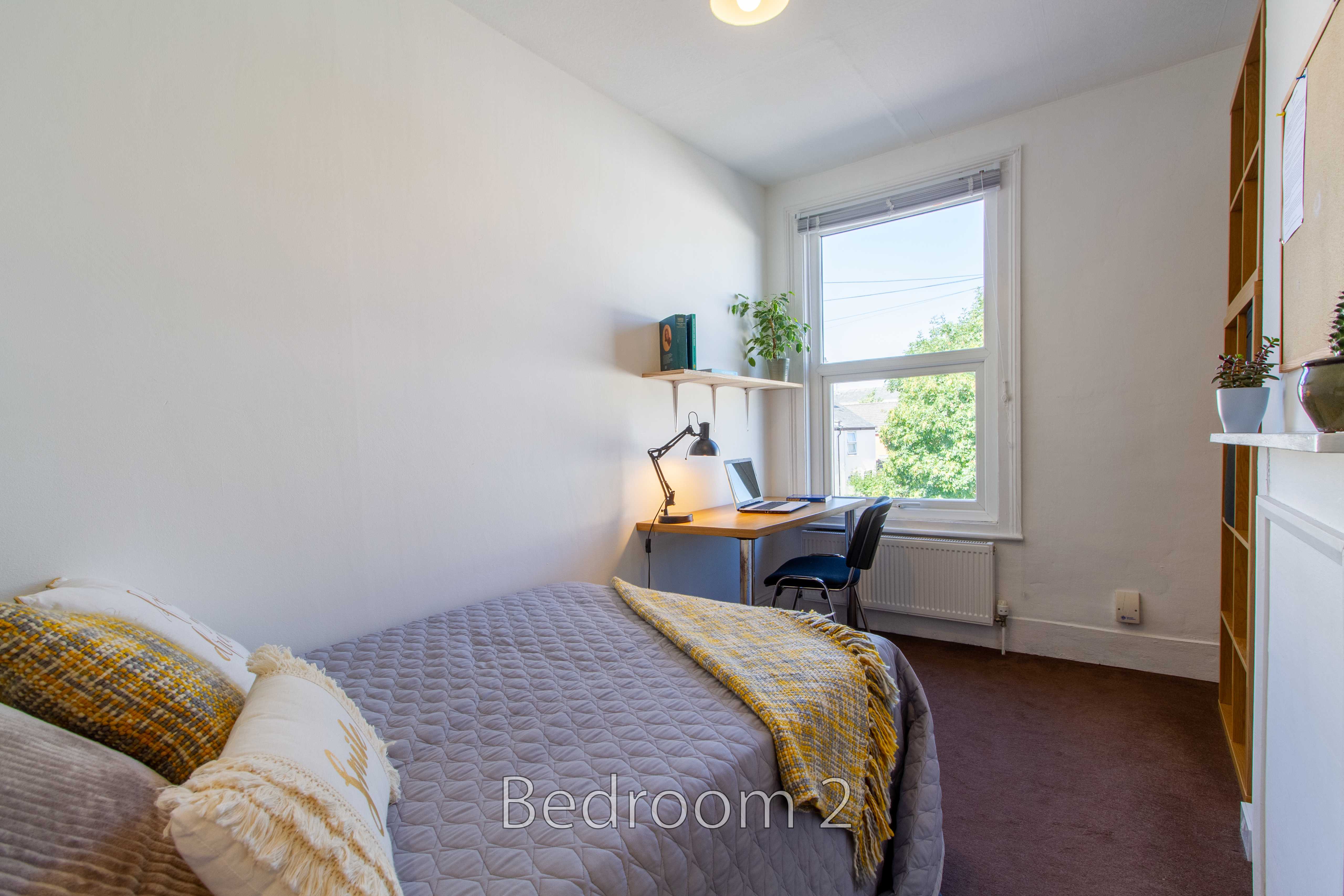 A furnished bedroom with a single bed, desk, chair, window, radiator and shelving.