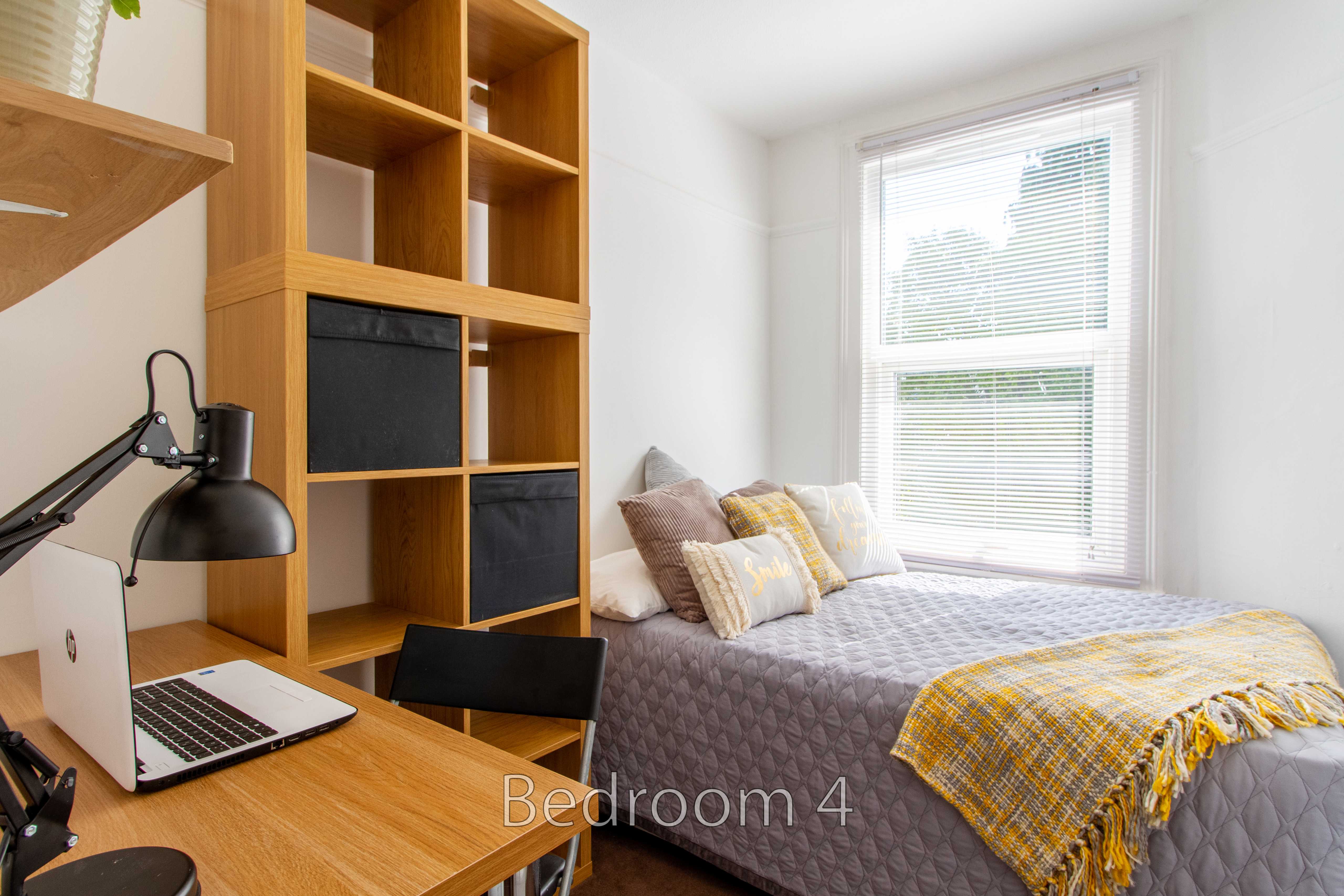A furnished bedroom with a single bed, desk, laptop, chair, shelving and a window.