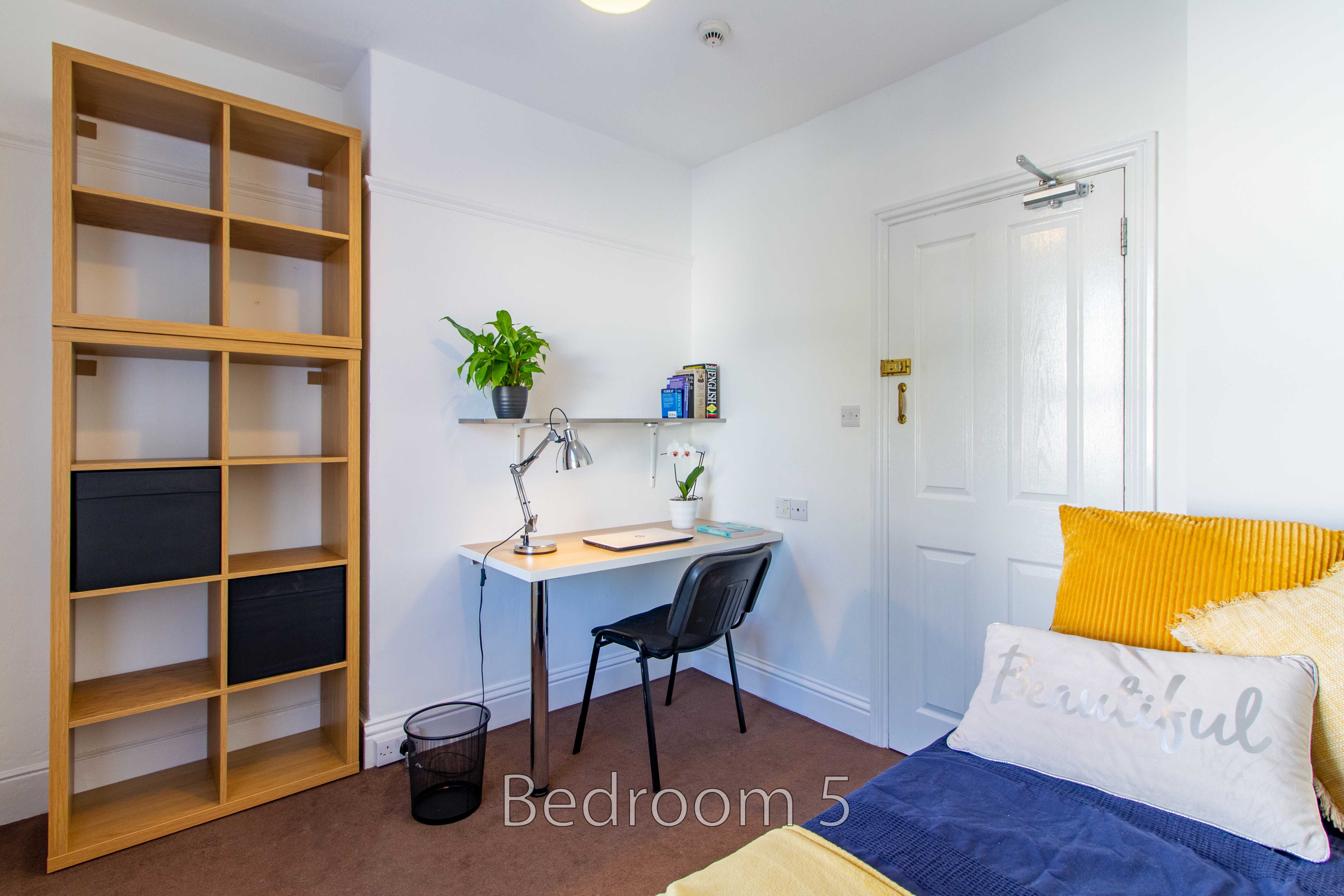 A furnished bedroom with a single bed, desk, chair, shelving unit, and white door.