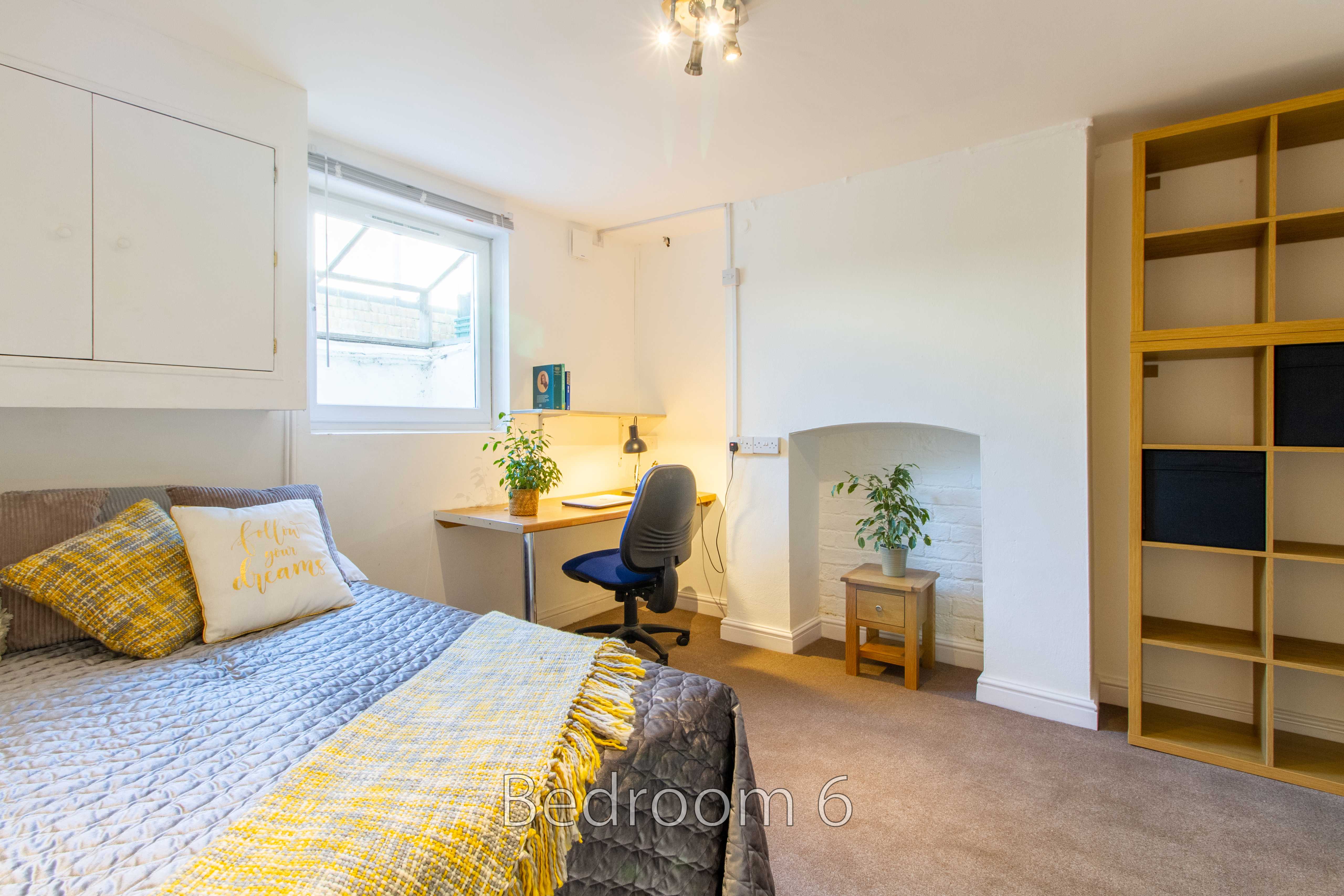 A furnished bedroom with a bed, desk and chair, fireplace, window, and open shelving.