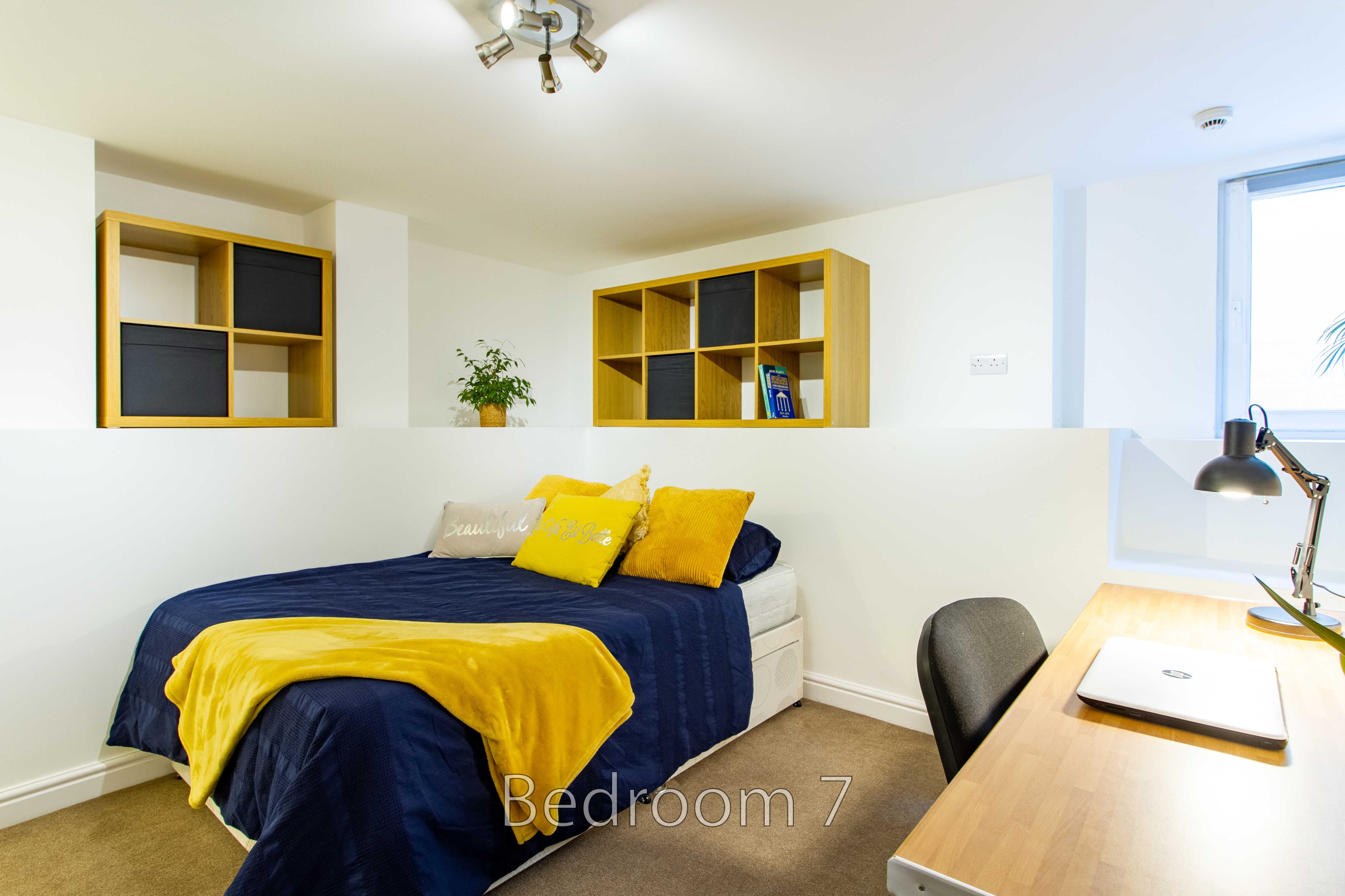 A furnished bedroom with a bed, colorful pillows, wall shelving, and a desk beside a window.