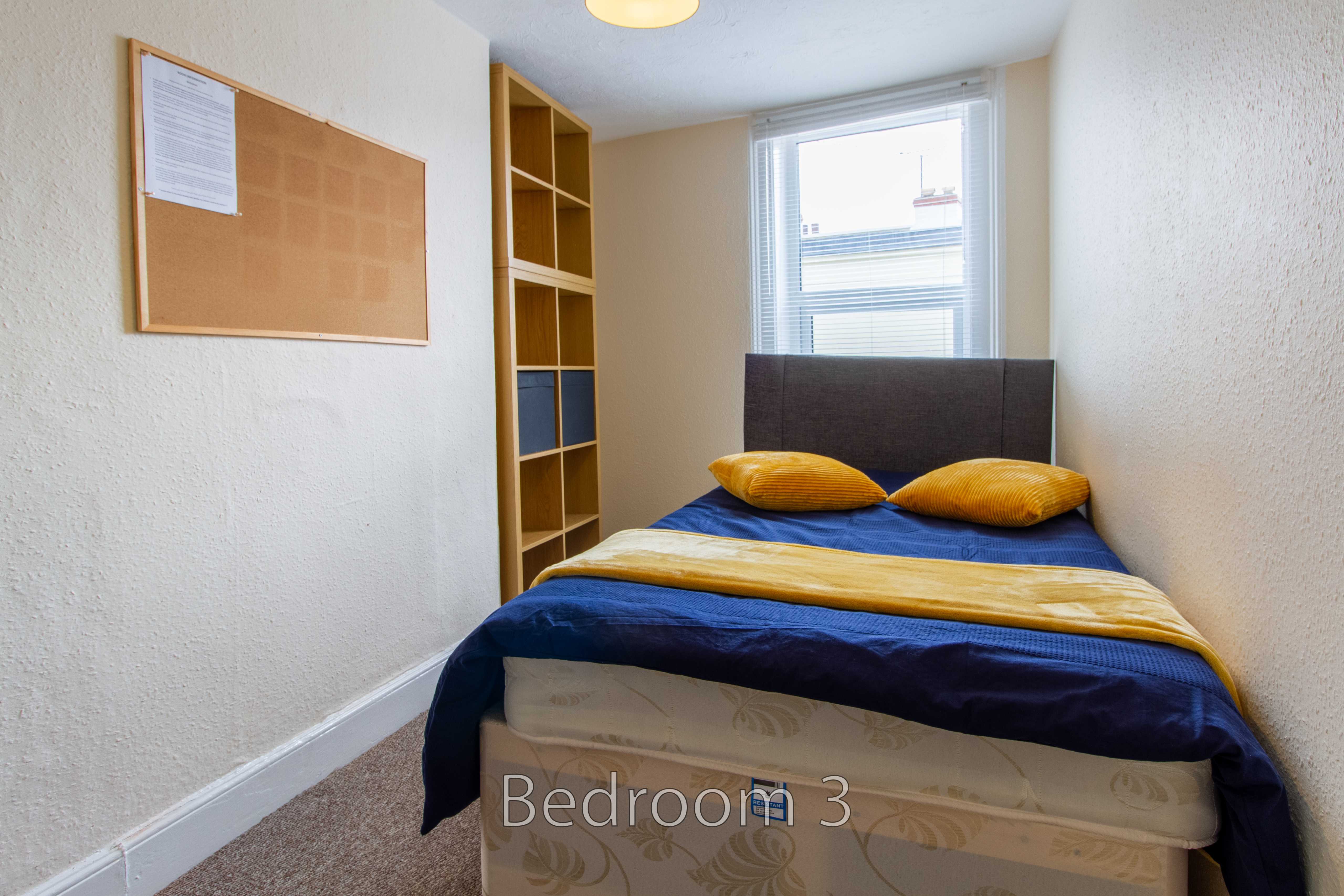 A small bedroom with a double bed, shelving, noticeboard and window.