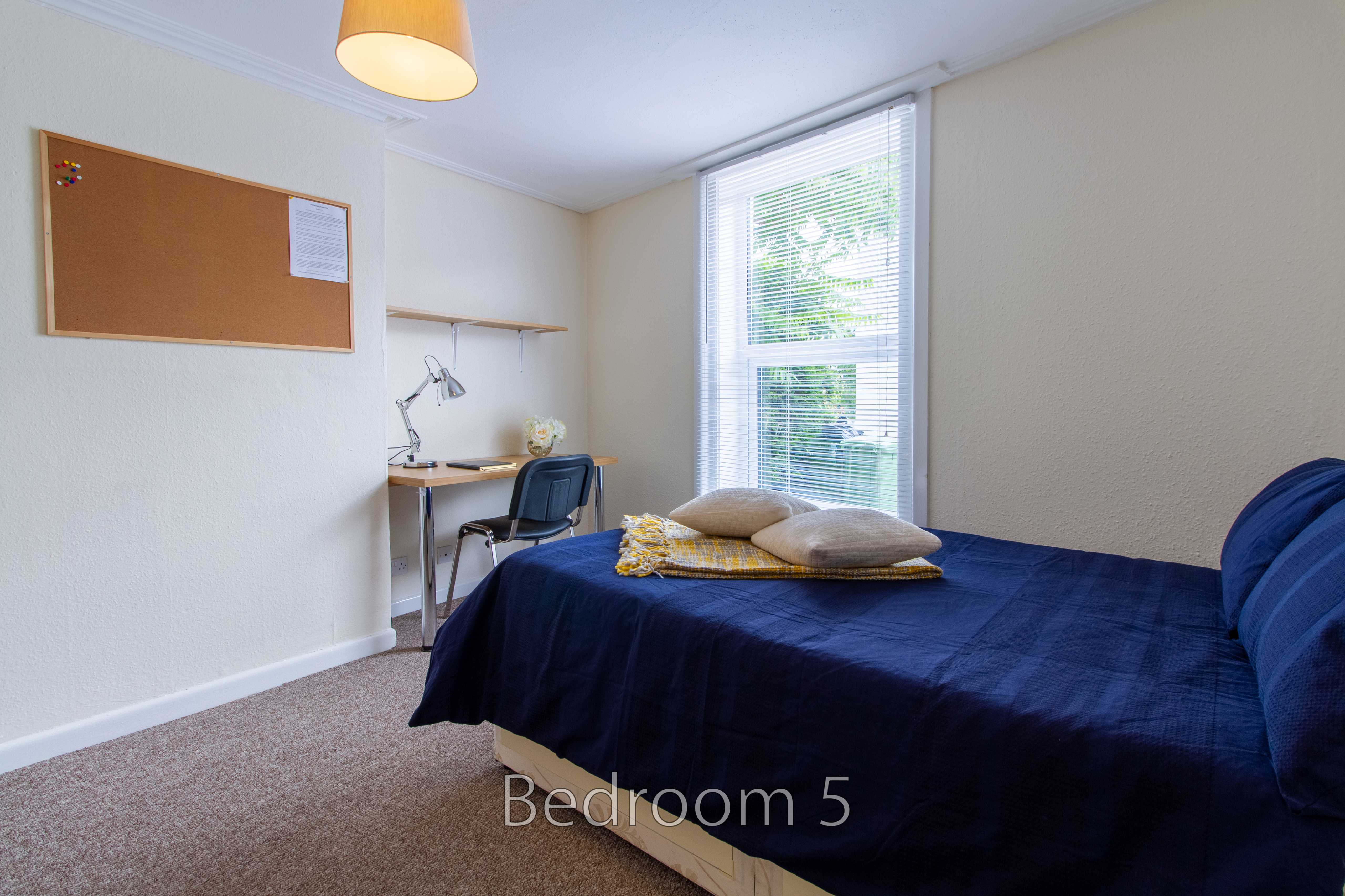 A furnished bedroom with a bed, desk, chair, window blinds and wall noticeboard.
