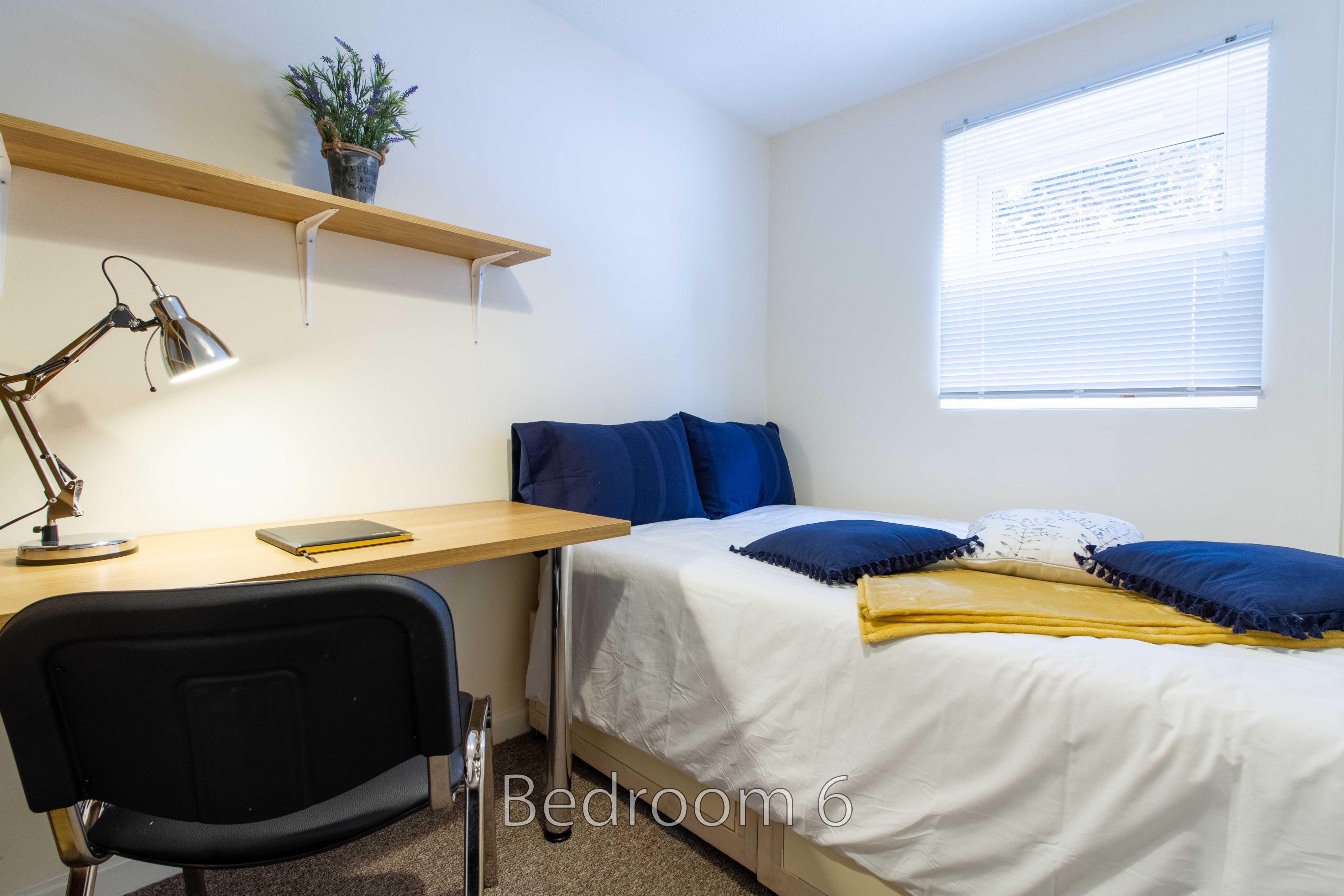 A tidy bedroom with a single bed, desk, chair, shelves, lamp and window blinds.