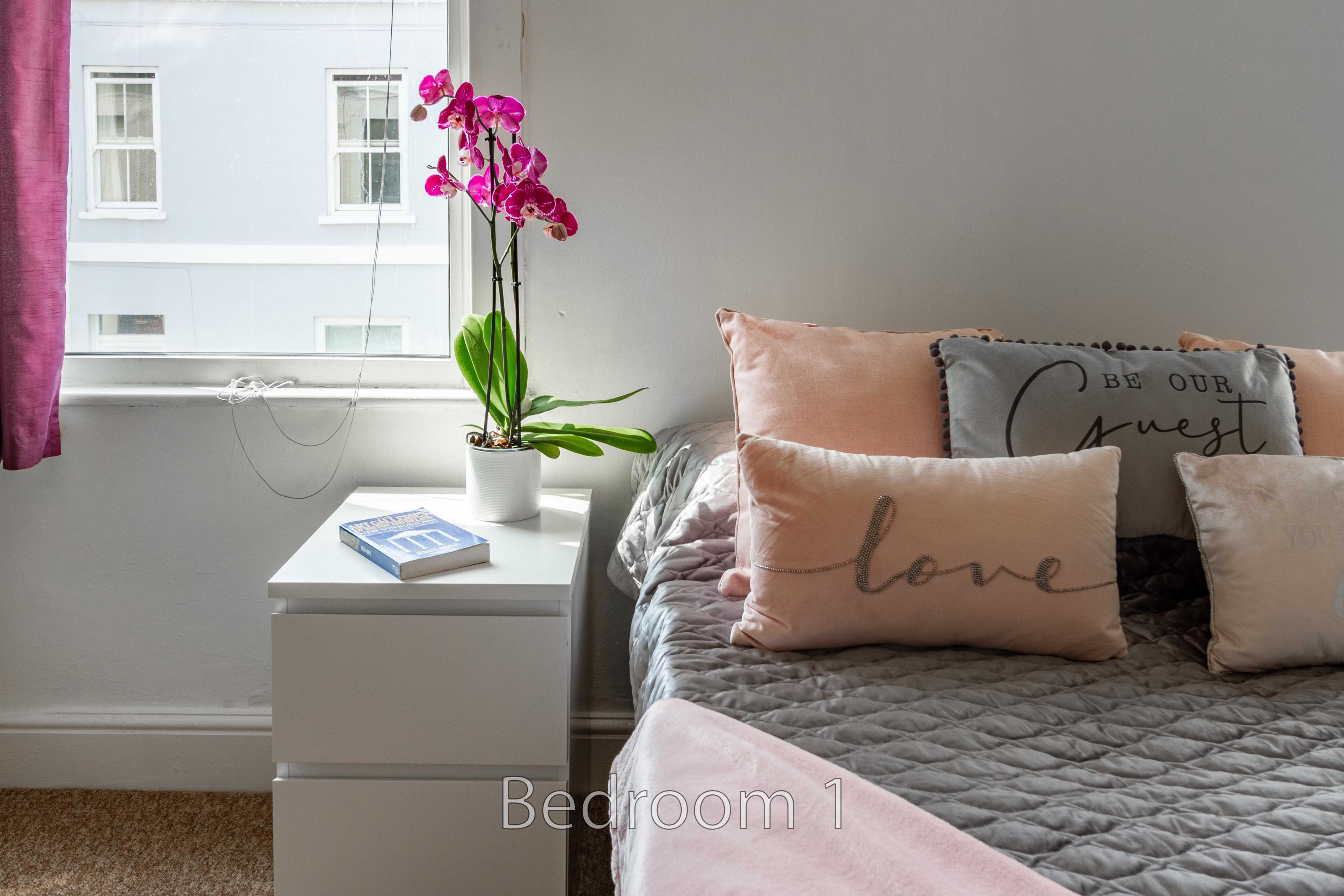 A bedroom with a neatly made bed, pink cushions, a bedside cabinet, an orchid, and a window.