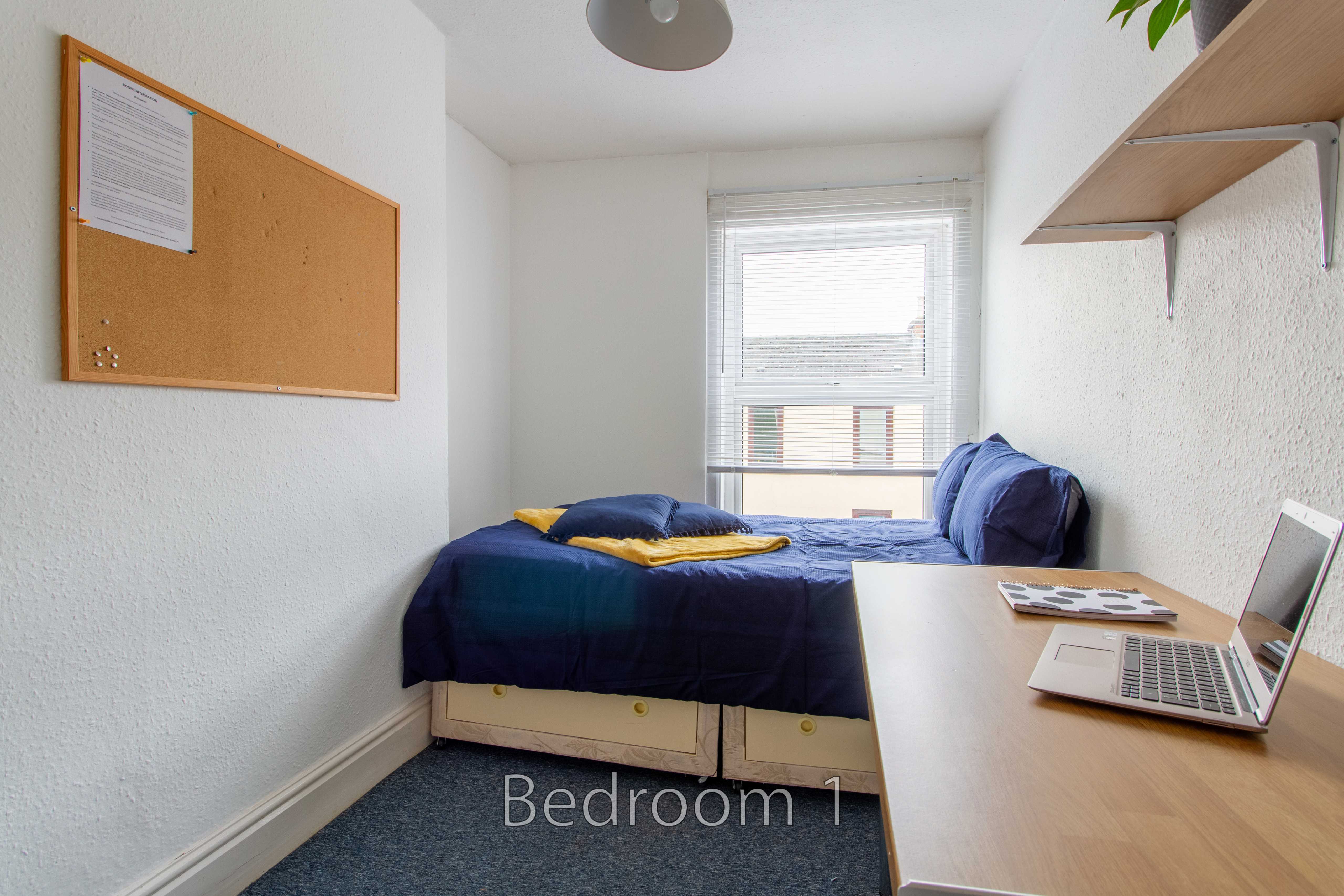 A small bedroom with a single bed, desk, laptop, window and wall-mounted shelves.