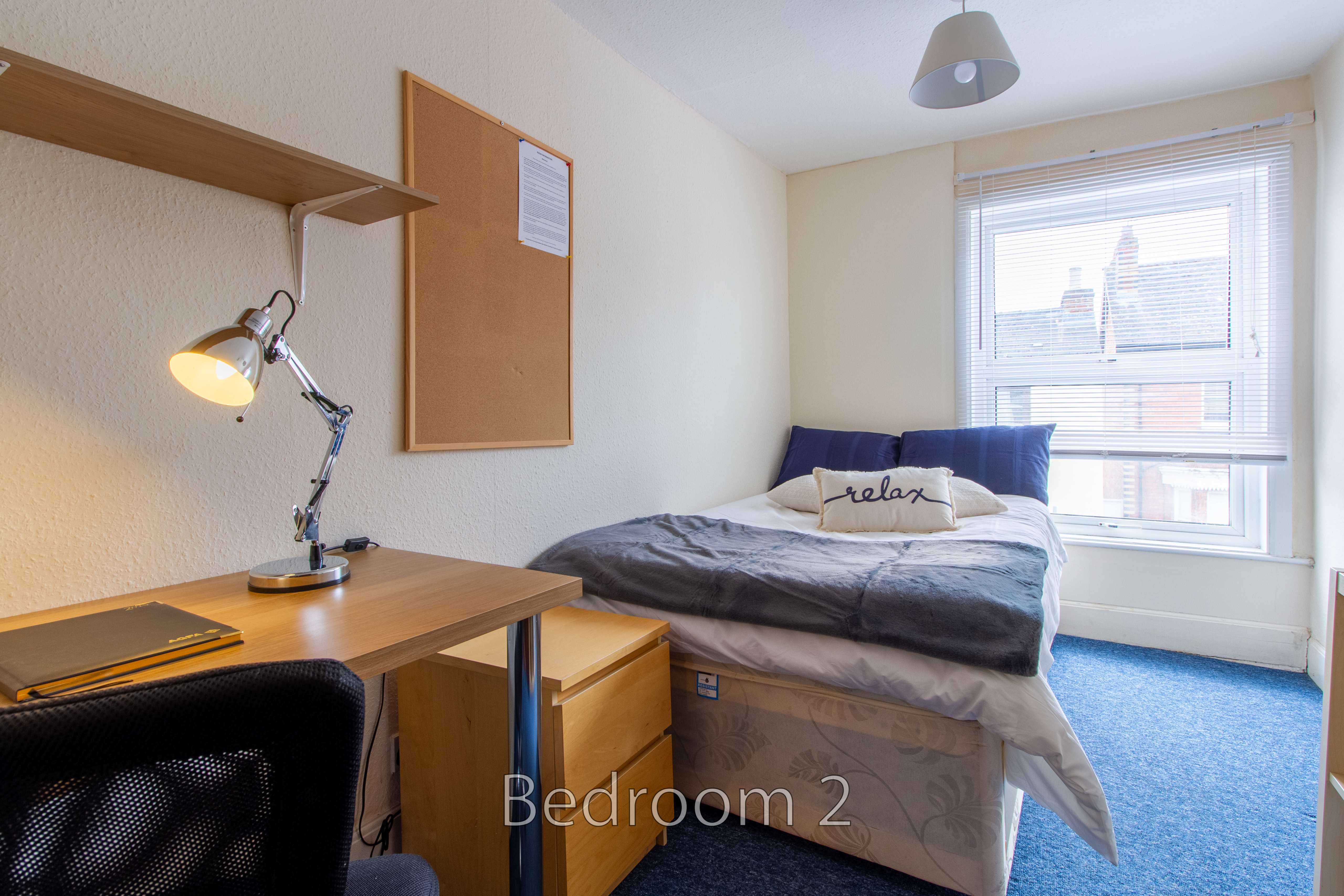 A furnished bedroom with a bed, desk, chair, shelves, noticeboard, lamp and window with blinds.