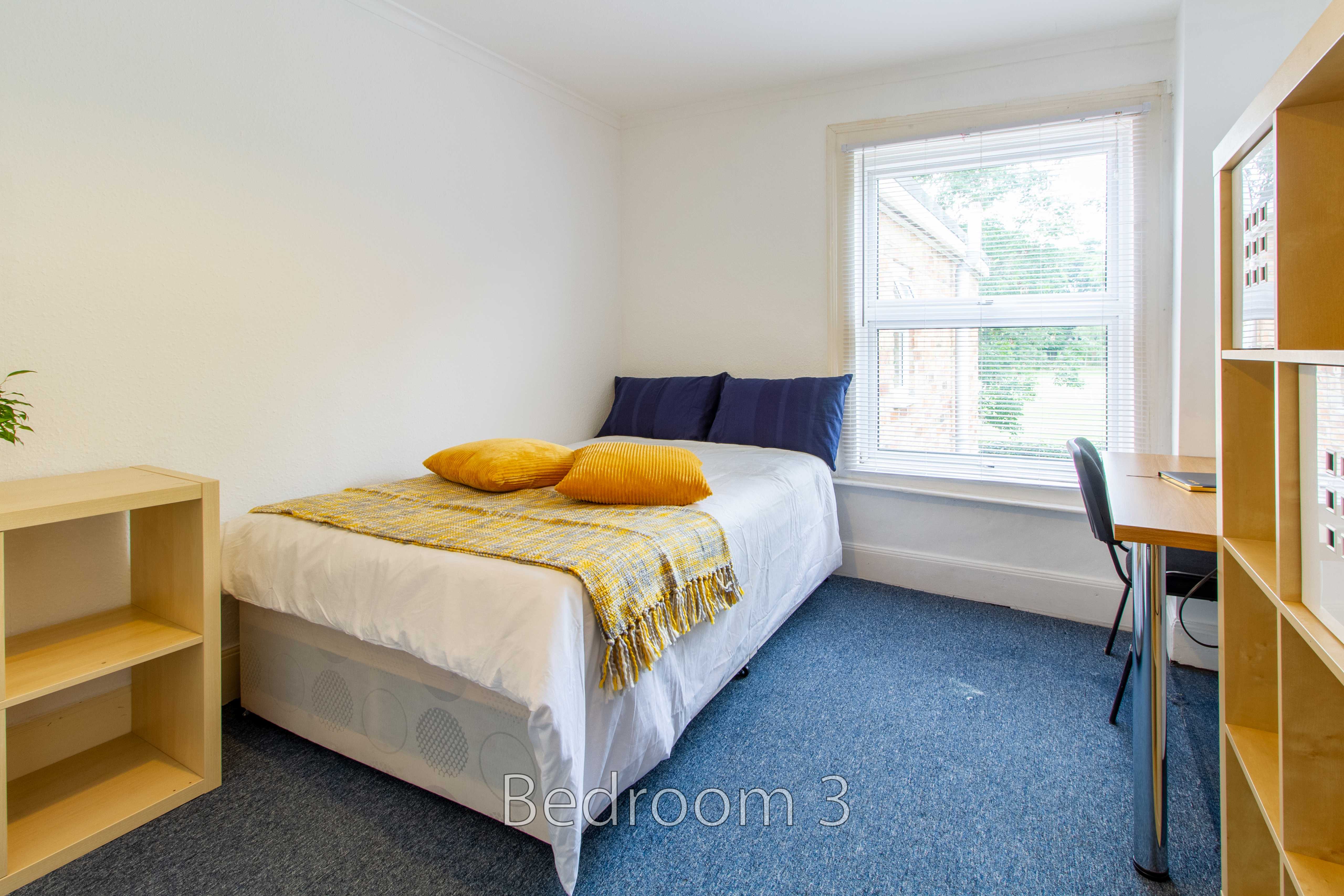 A furnished bedroom with a bed, desk, shelving, window and blue carpet.