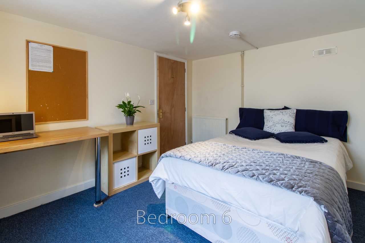 A furnished bedroom with a double bed, blue carpet, desk, shelving and a wall-mounted noticeboard.