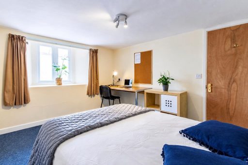 A bedroom with a bed, desk, chair, window, curtains, storage unit and wooden door.