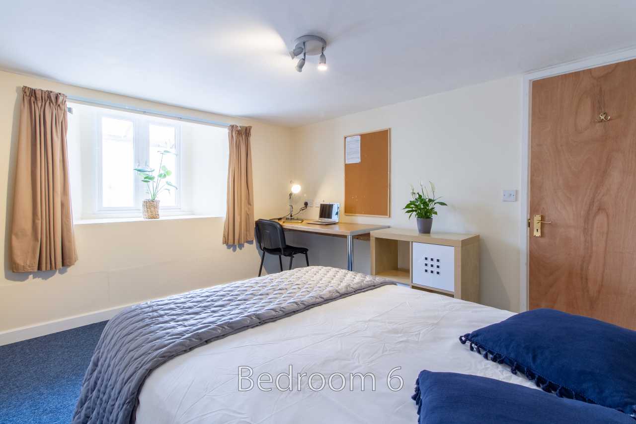 A bedroom with a bed, desk, chair, window, curtains, storage unit and wooden door.