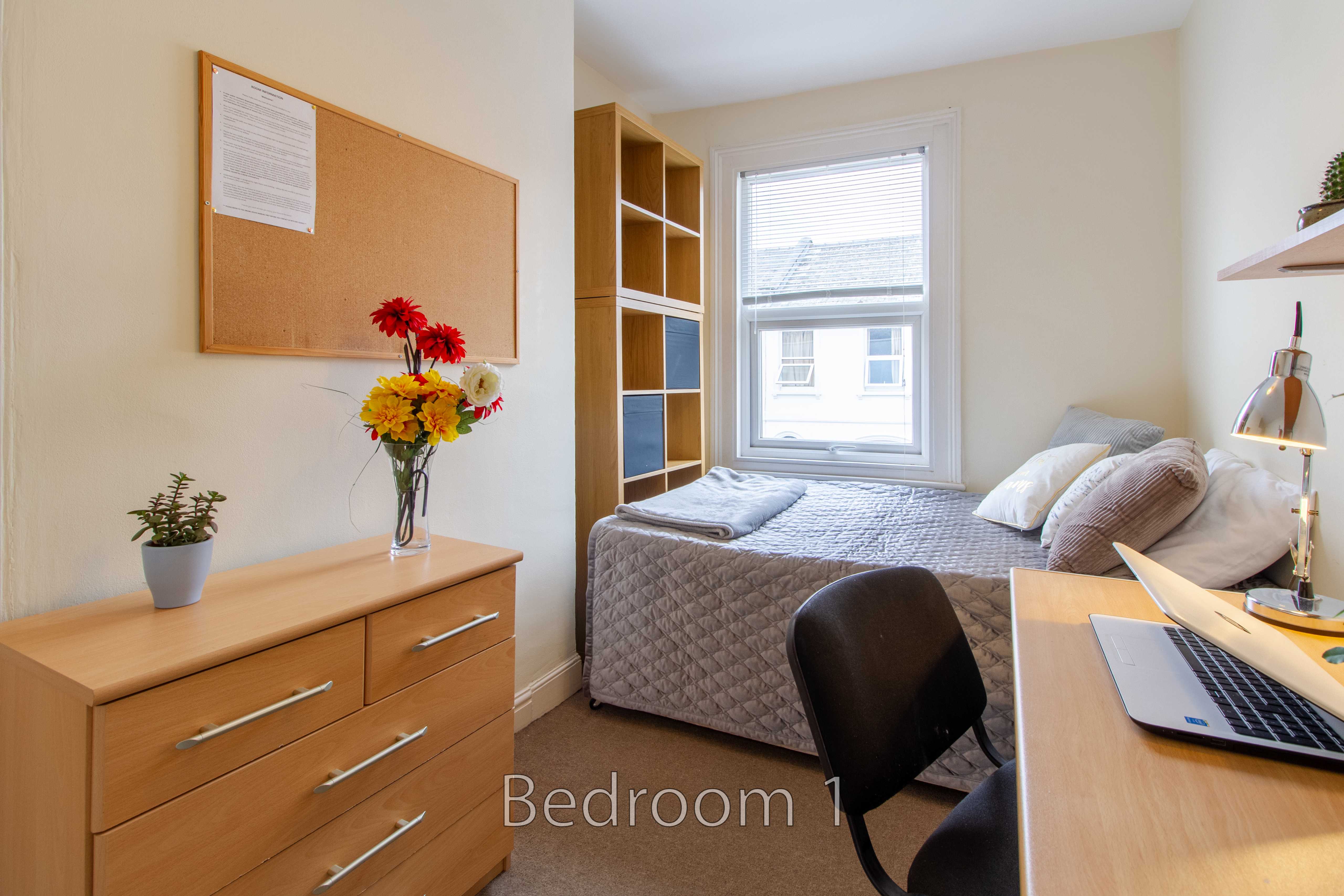 Furnished bedroom with a single bed, desk, chair, chest of drawers, shelving and a window.