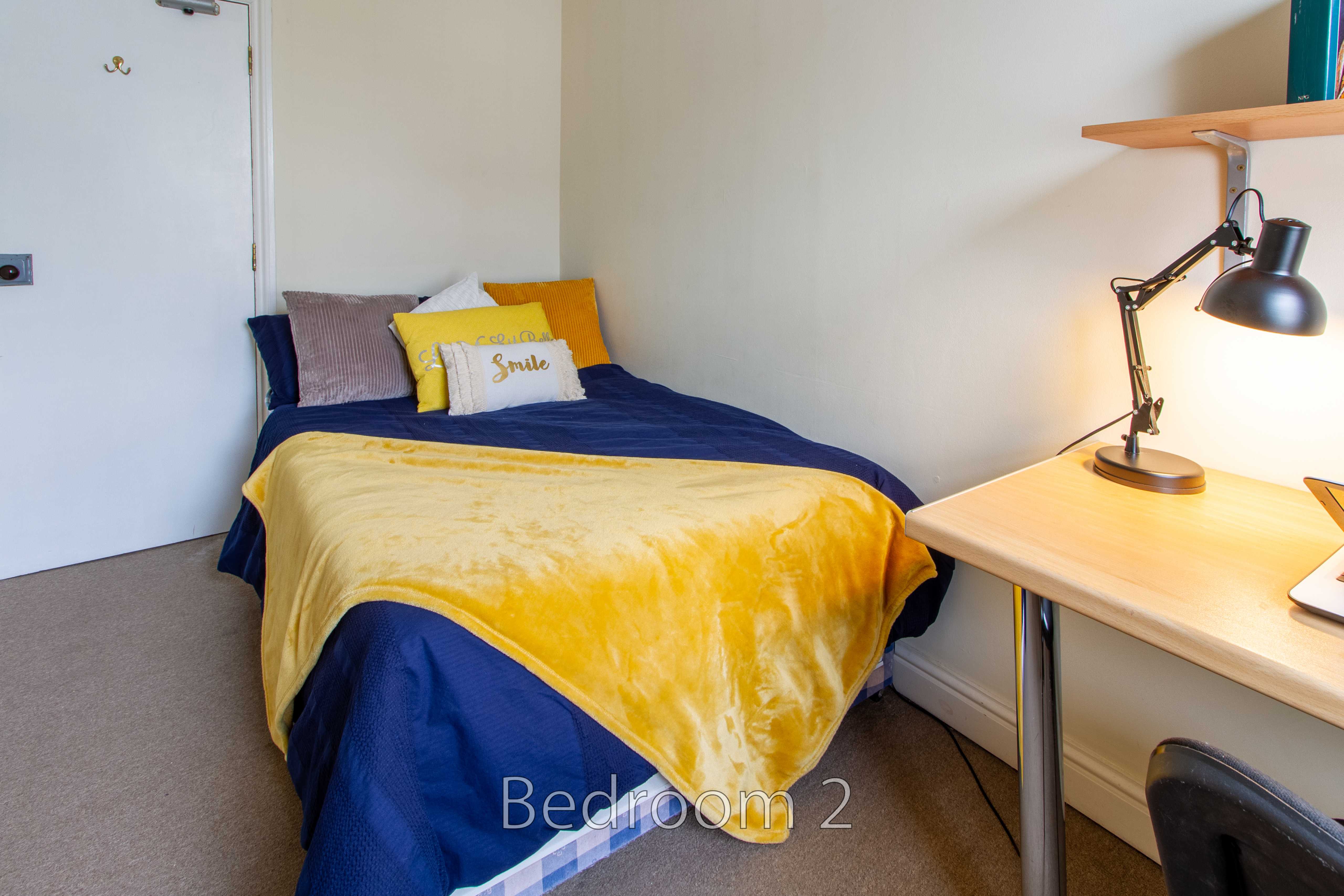 A bedroom with a single bed, yellow and blue bedding, a desk, chair and lamp.