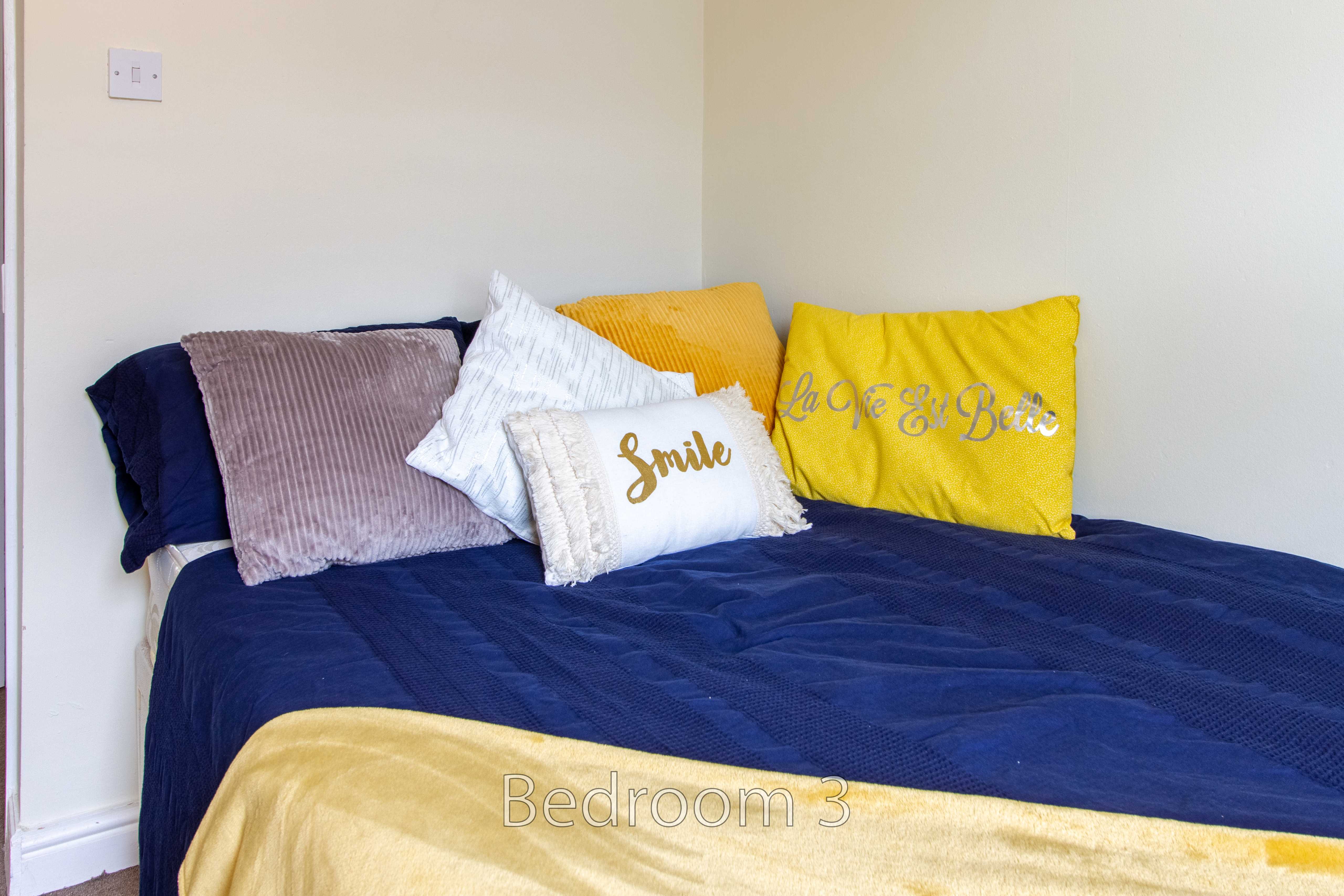 A bed with dark blue bedding, yellow and white pillows, and a yellow throw against a plain wall.