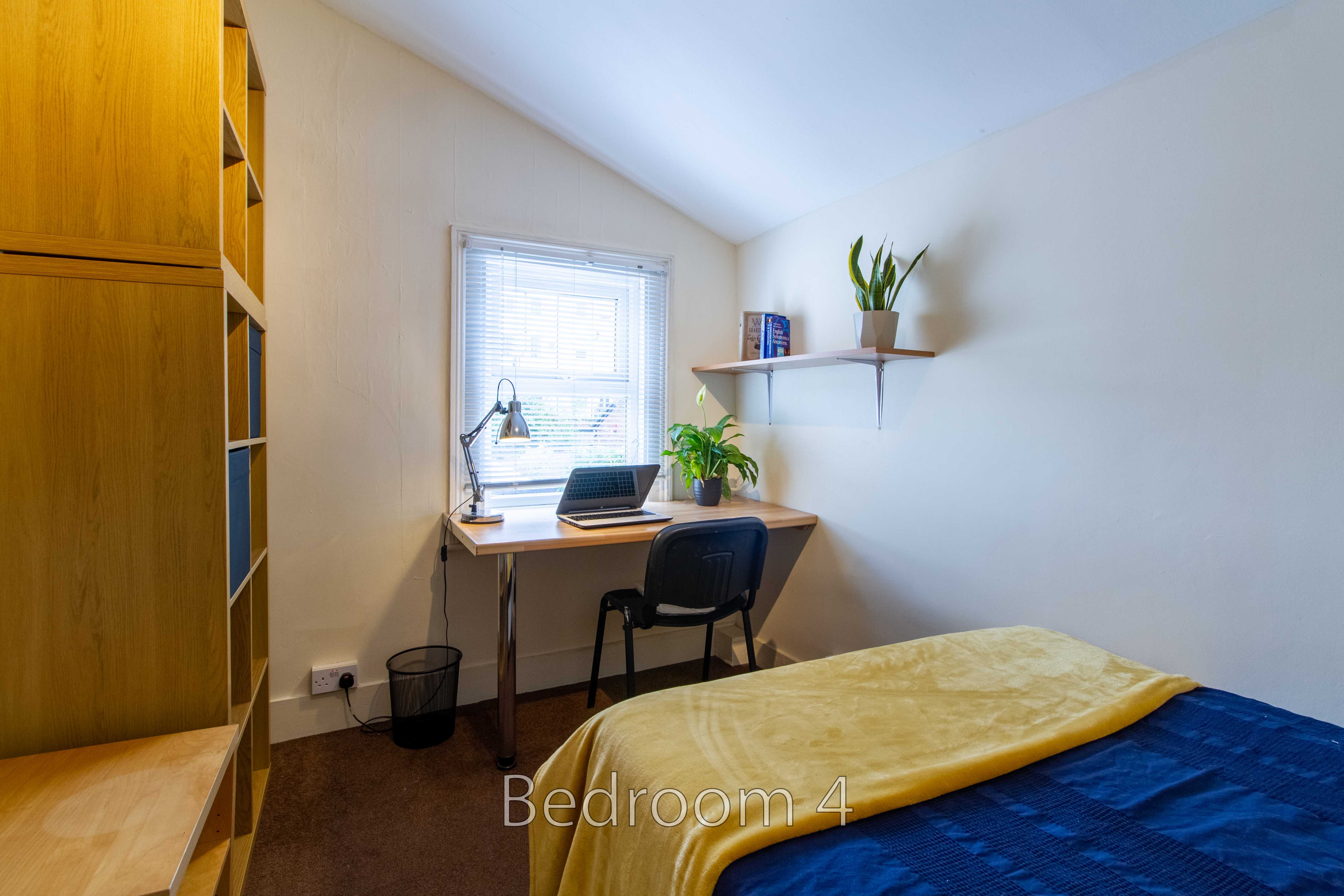 A small bedroom with a single bed, desk, chair, window with blinds, shelving, and a wardrobe.