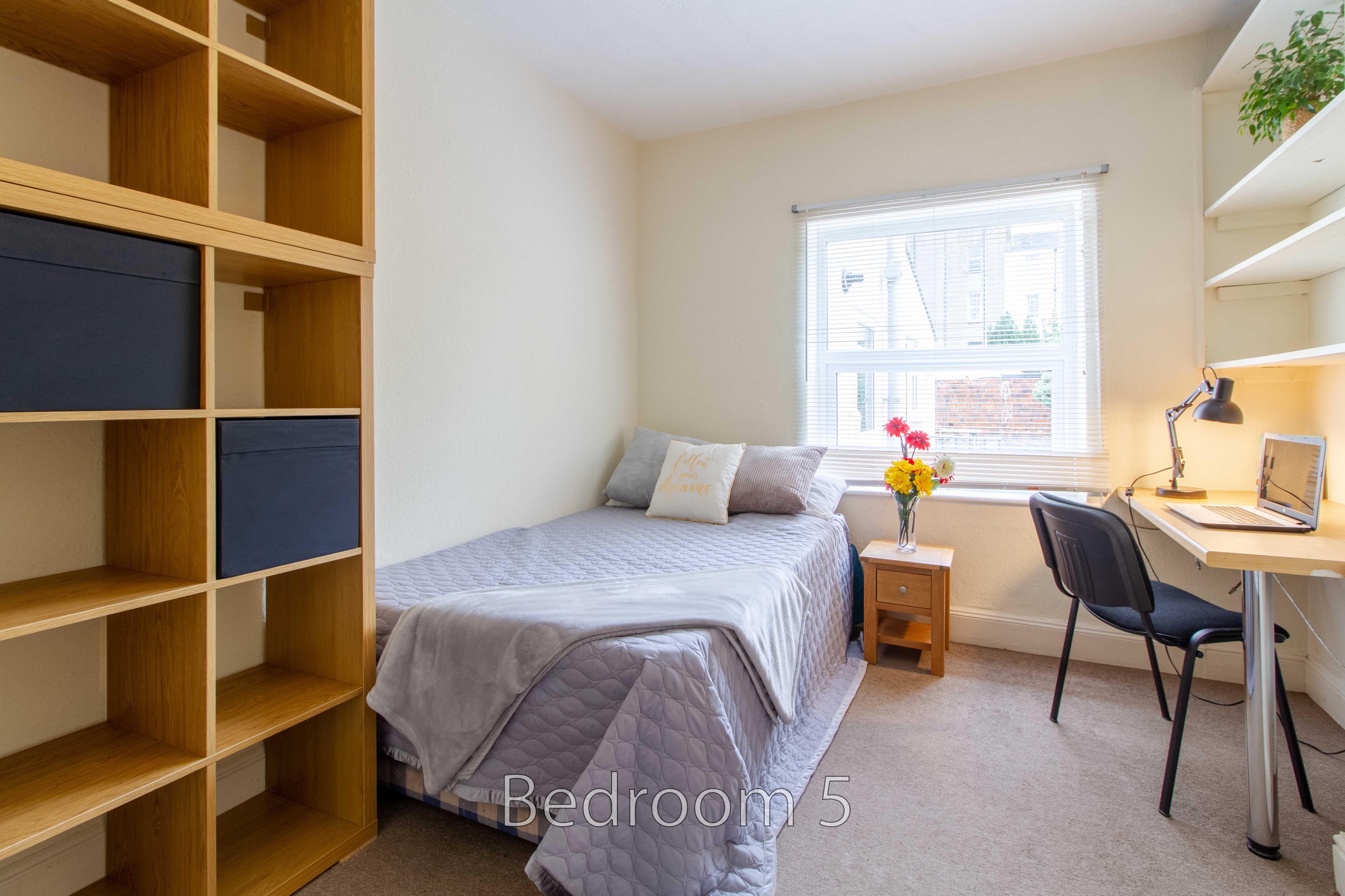A single bedroom with a bed, desk, chair, shelving and a window.