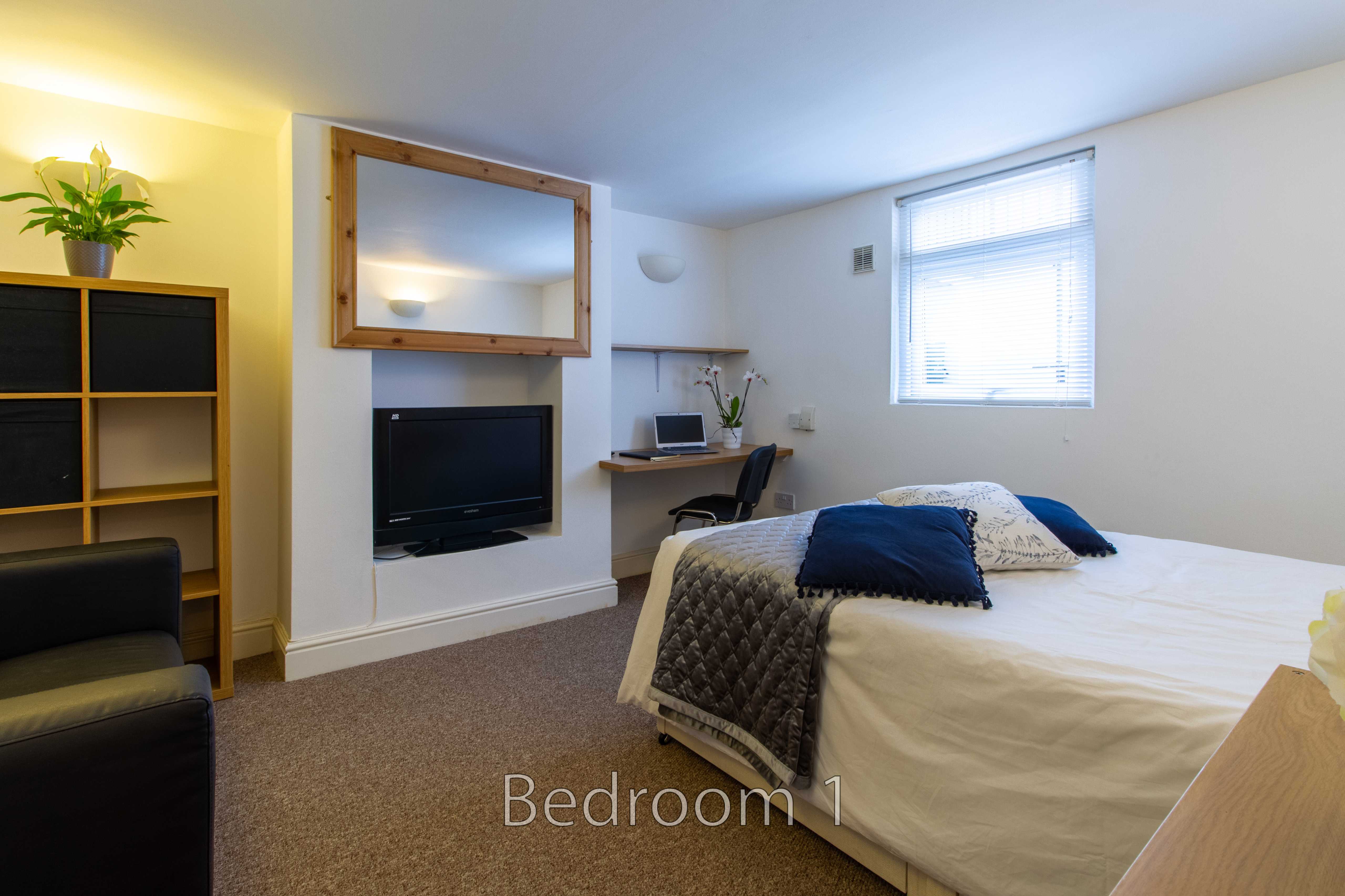 A furnished bedroom with a double bed, television, desk, shelving and a window with blinds.