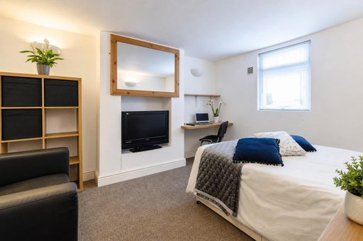 A furnished bedroom with a double bed, television, desk, shelving and a window with blinds.