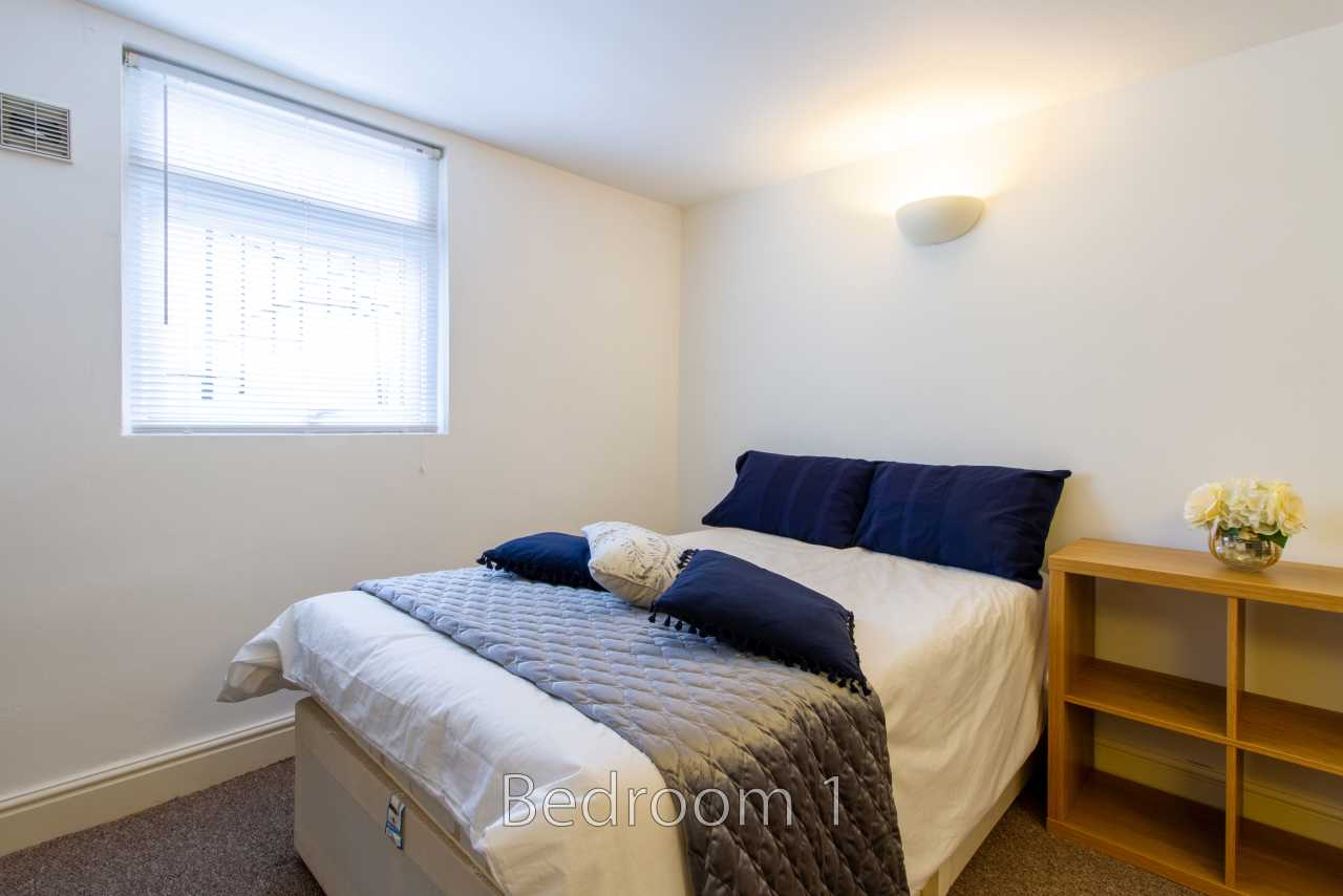 A furnished bedroom with a double bed, window blinds, wall light and wooden shelving.
