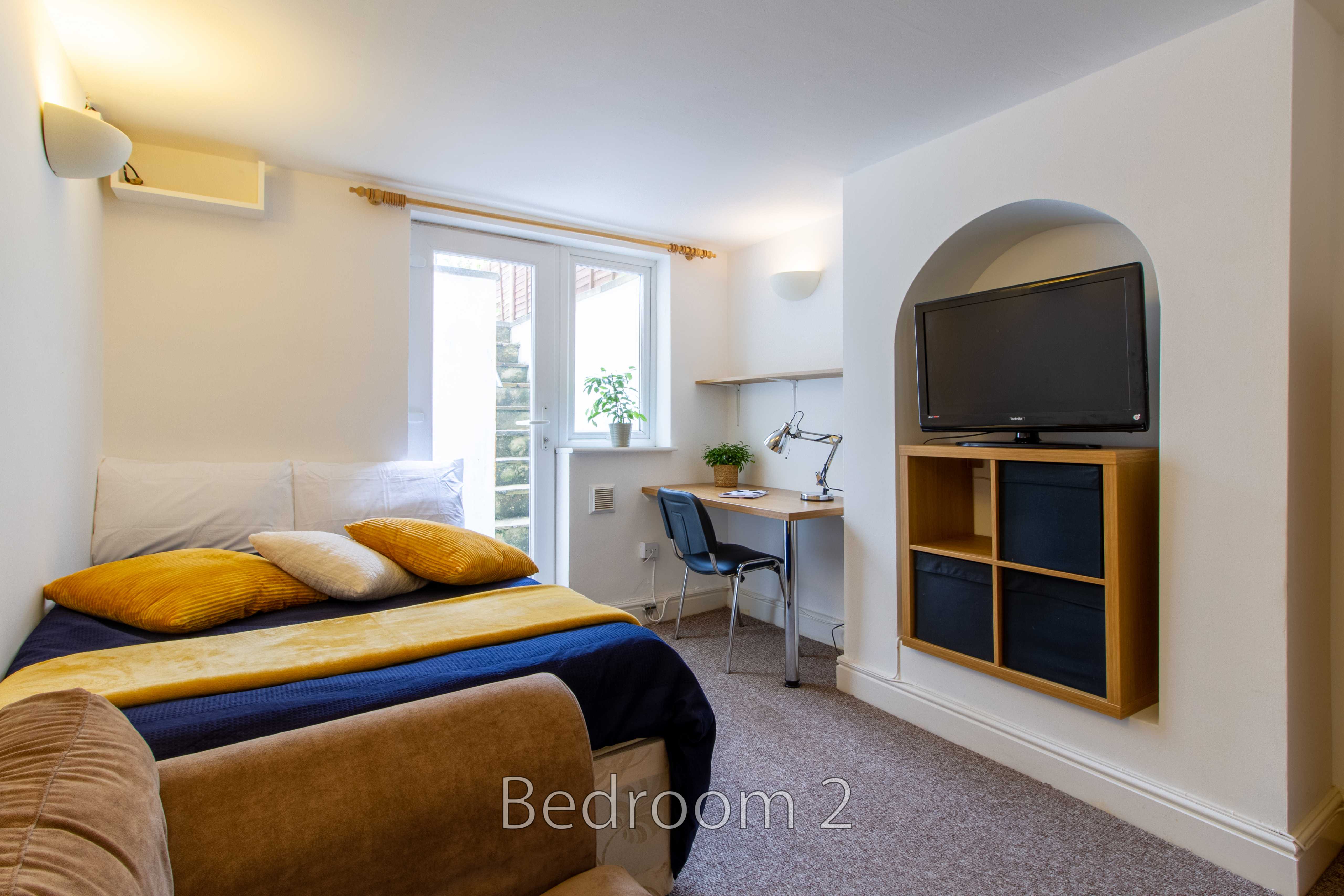 A furnished bedroom with a bed, window, desk and chair, wall-mounted television, and shelving.