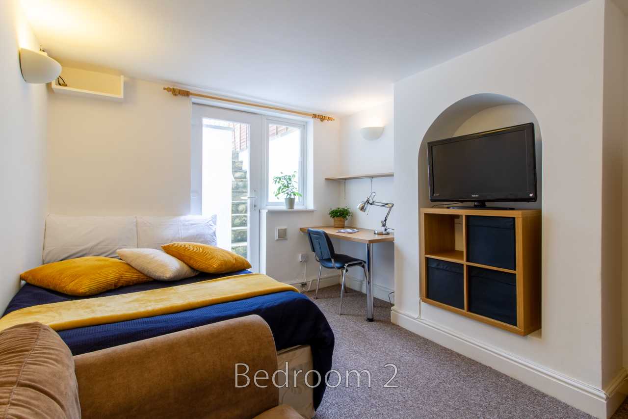 A furnished bedroom with a bed, window, desk and chair, wall-mounted television, and shelving.