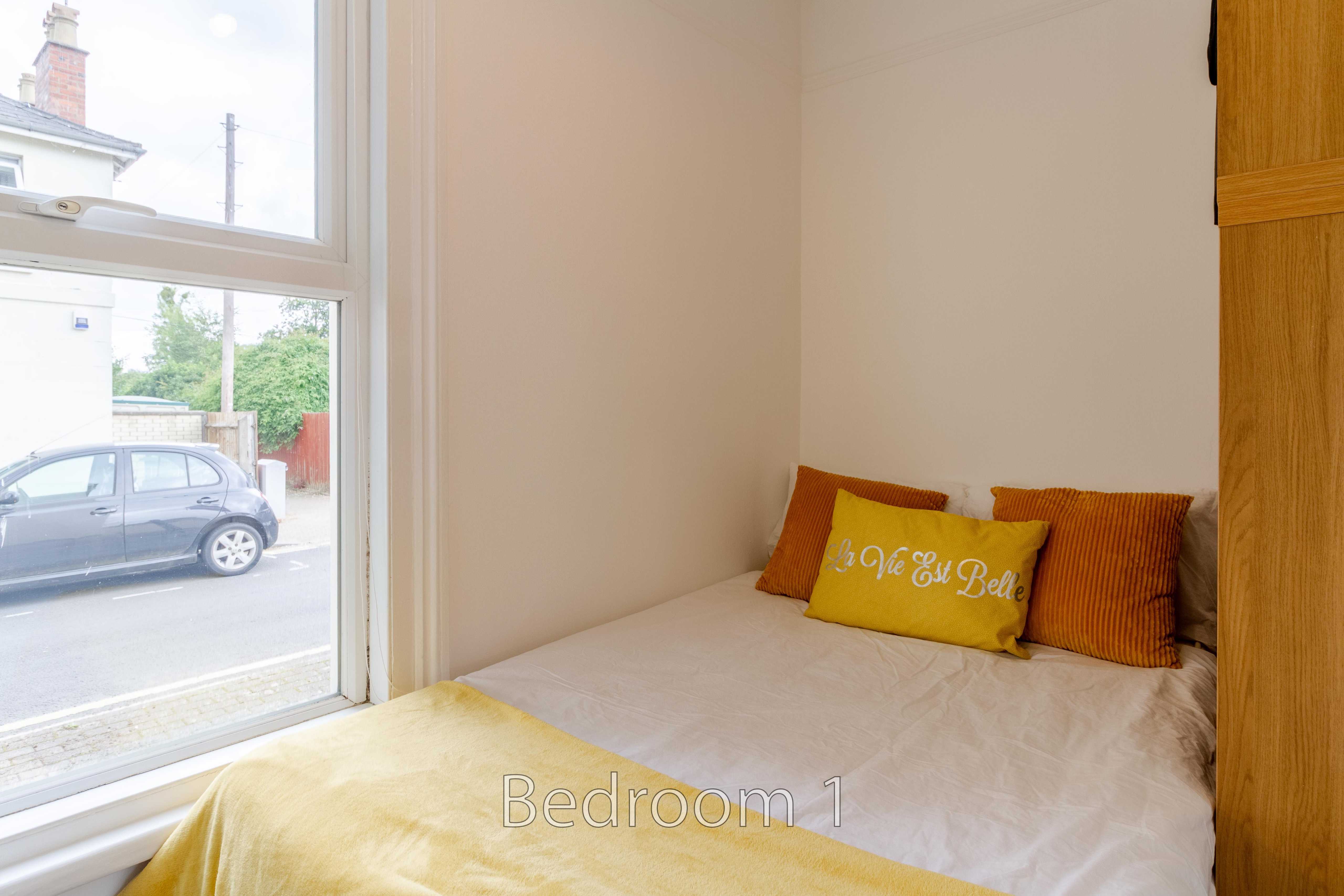 A small bedroom with a single bed, yellow and orange cushions, a large window, and a wooden wardrobe.